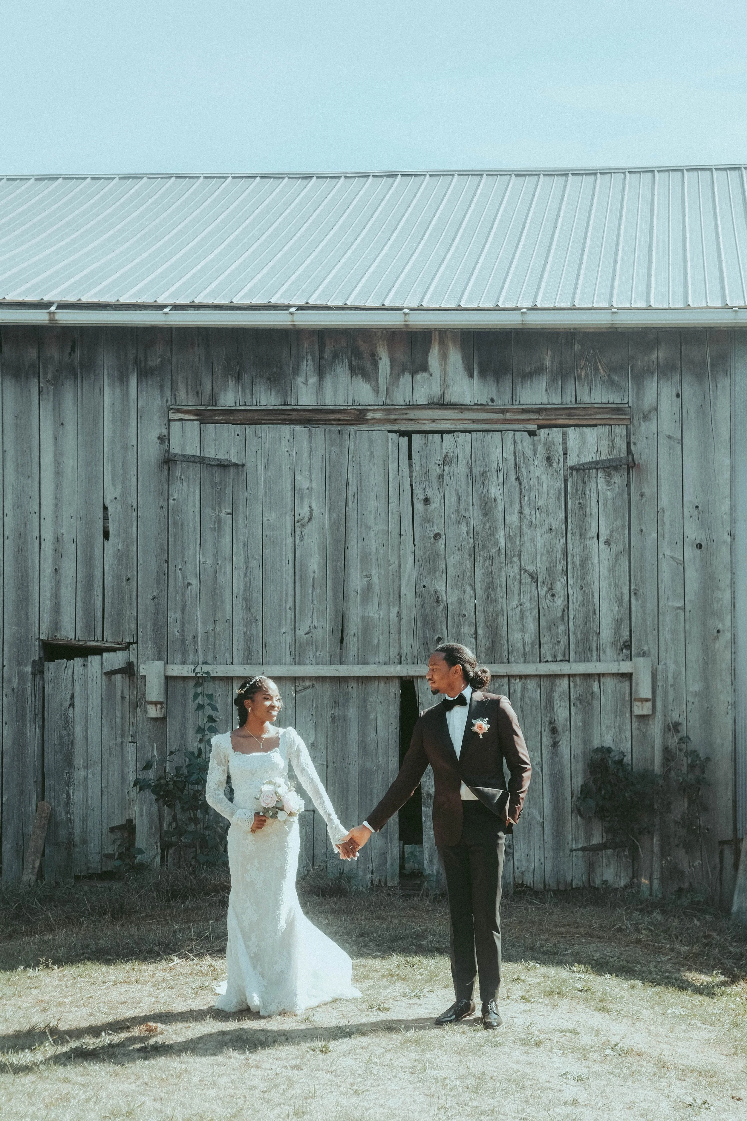 A bride and groom holding hands outdoors in front of a weathered wooden barn during their wedding. The bride is in a white lace wedding gown holding a bouquet, and the groom is in a black tuxedo with a bow tie.