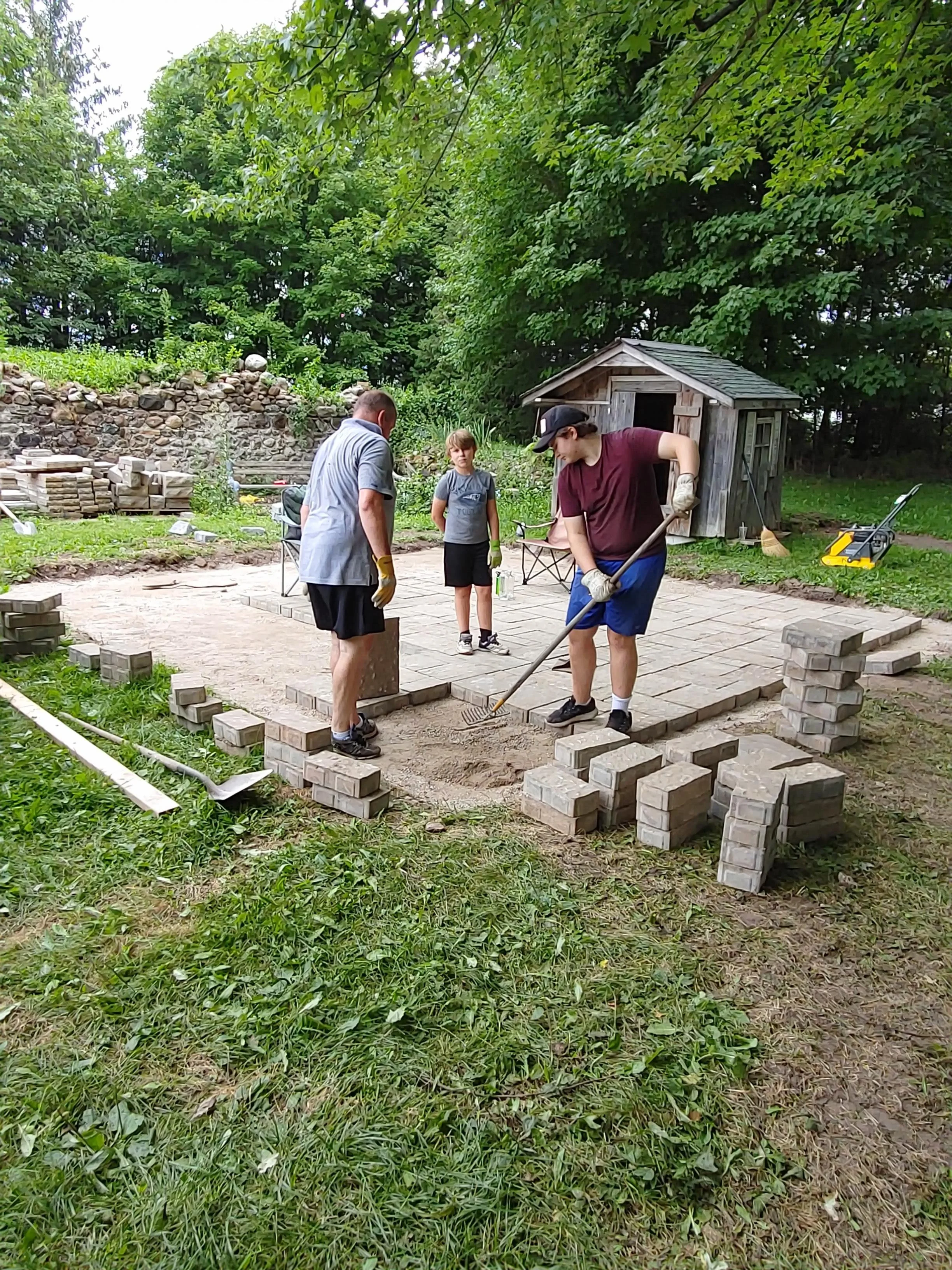 People laying bricks to build a patio in a backyard with trees and a small wooden shed.