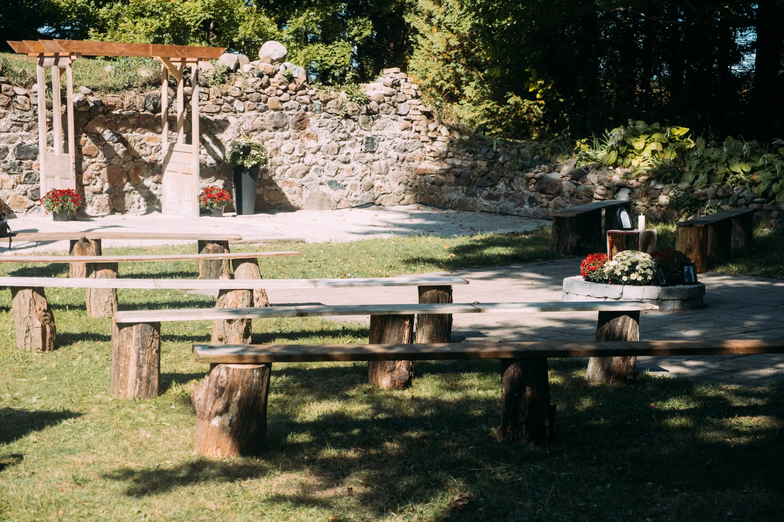 Outdoor memorial site with wooden benches, a stone platform with flowers and a candle, and a stone wall backdrop, set in a grassy area with trees.