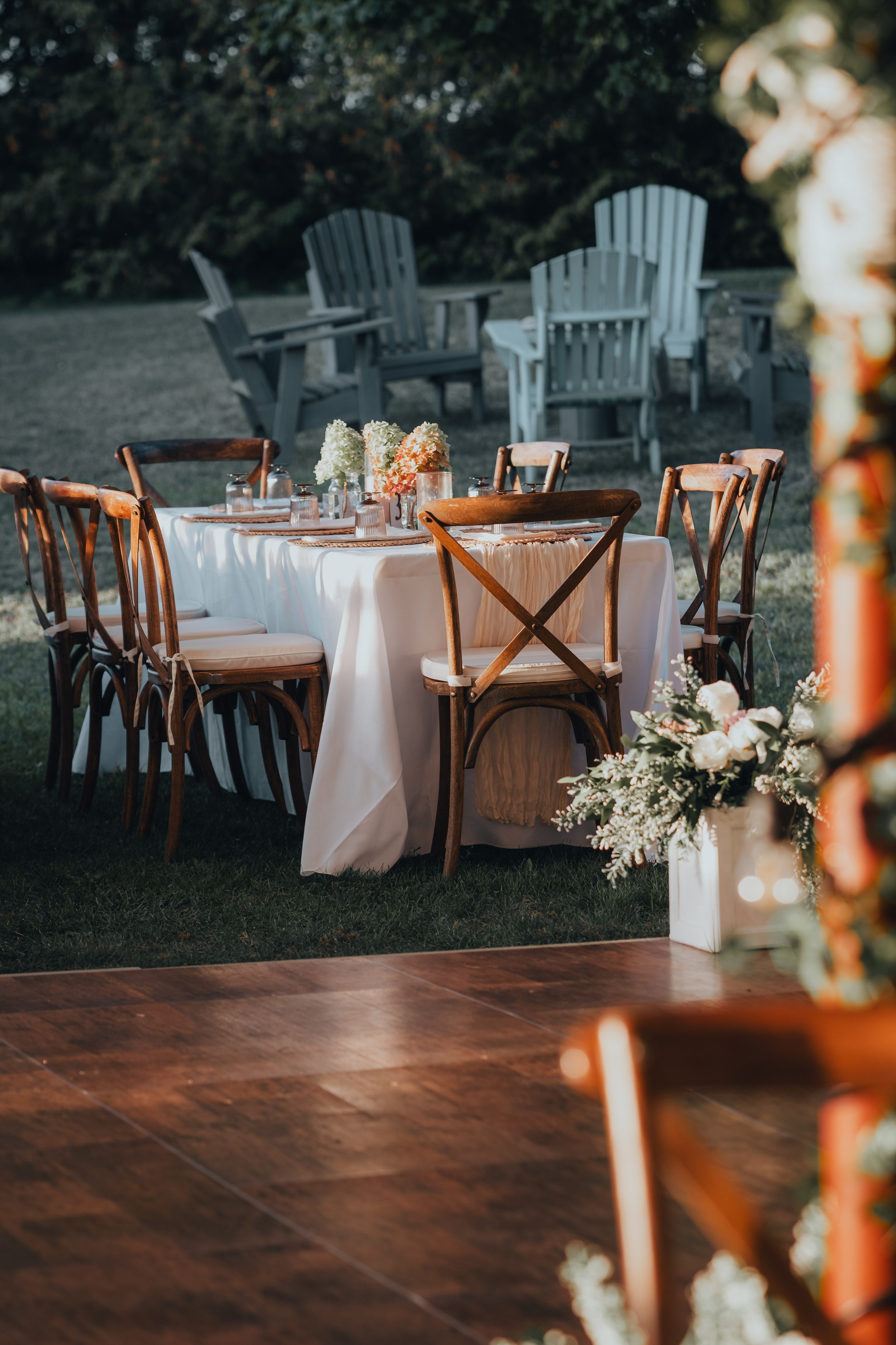 Elegant outdoor event setup with a long dining table covered with a white tablecloth, surrounded by wooden chairs, with floral centerpieces and glassware on the table. In the background, there are Adirondack chairs on a grassy area, and part of a dec