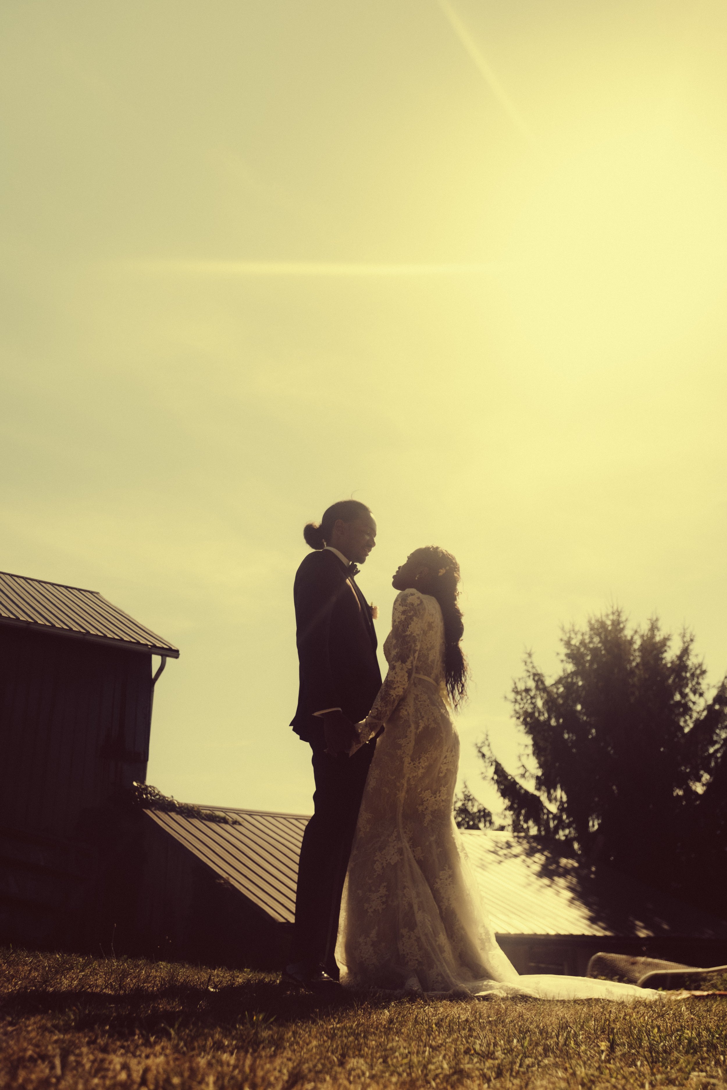 Silhouette of a bride and groom holding hands and facing each other outdoors during sunset, with a building and trees in the background.