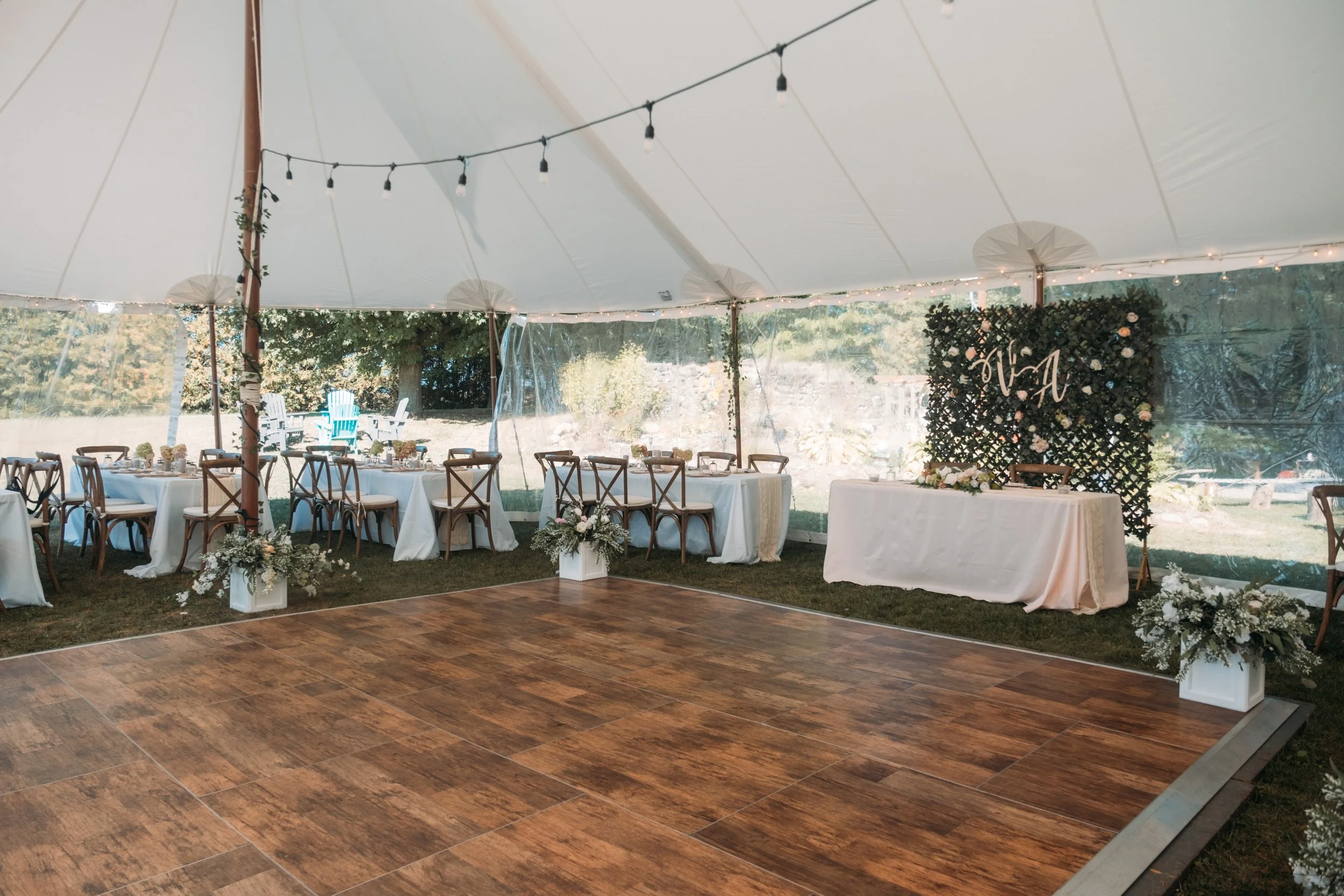 Empty wedding reception tent with a wooden dance floor, decorated with fairy lights, floral arrangements, and a sweetheart table with a floral backdrop.