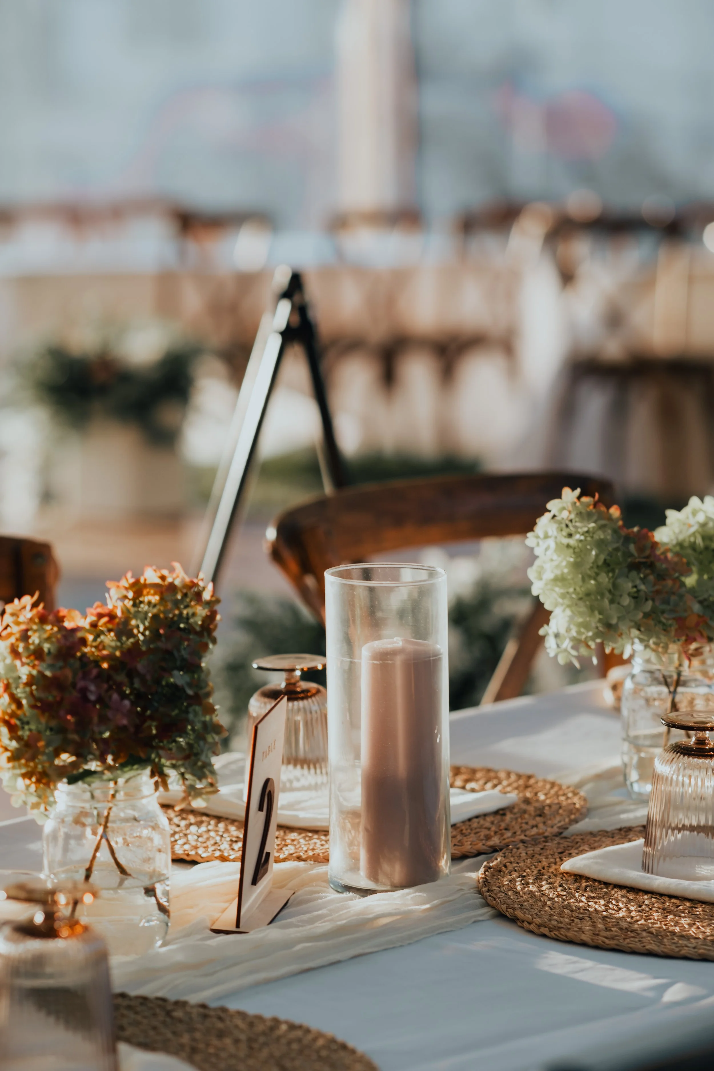 Decorated table with floral arrangements, candles, and table number at a sunny outdoor event.