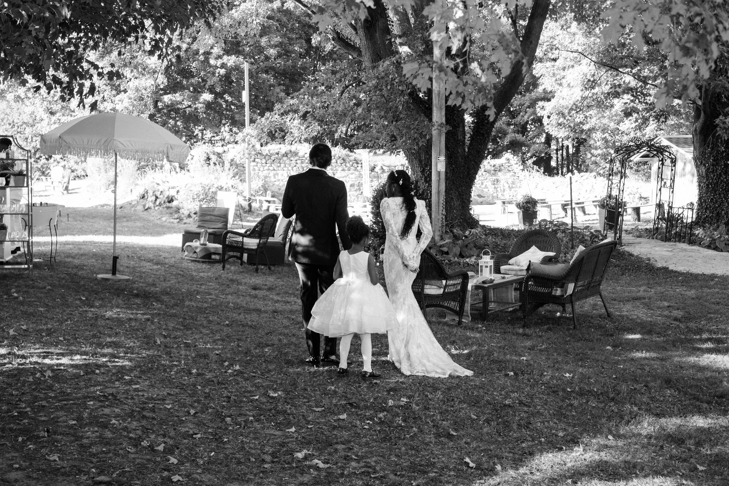A wedding scene in a garden with a bride in a long lace dress, a groom in a dark suit, and a young girl in a white dress standing beneath a large tree. There are outdoor chairs, a table, and decorative elements like lanterns and garden furniture surr