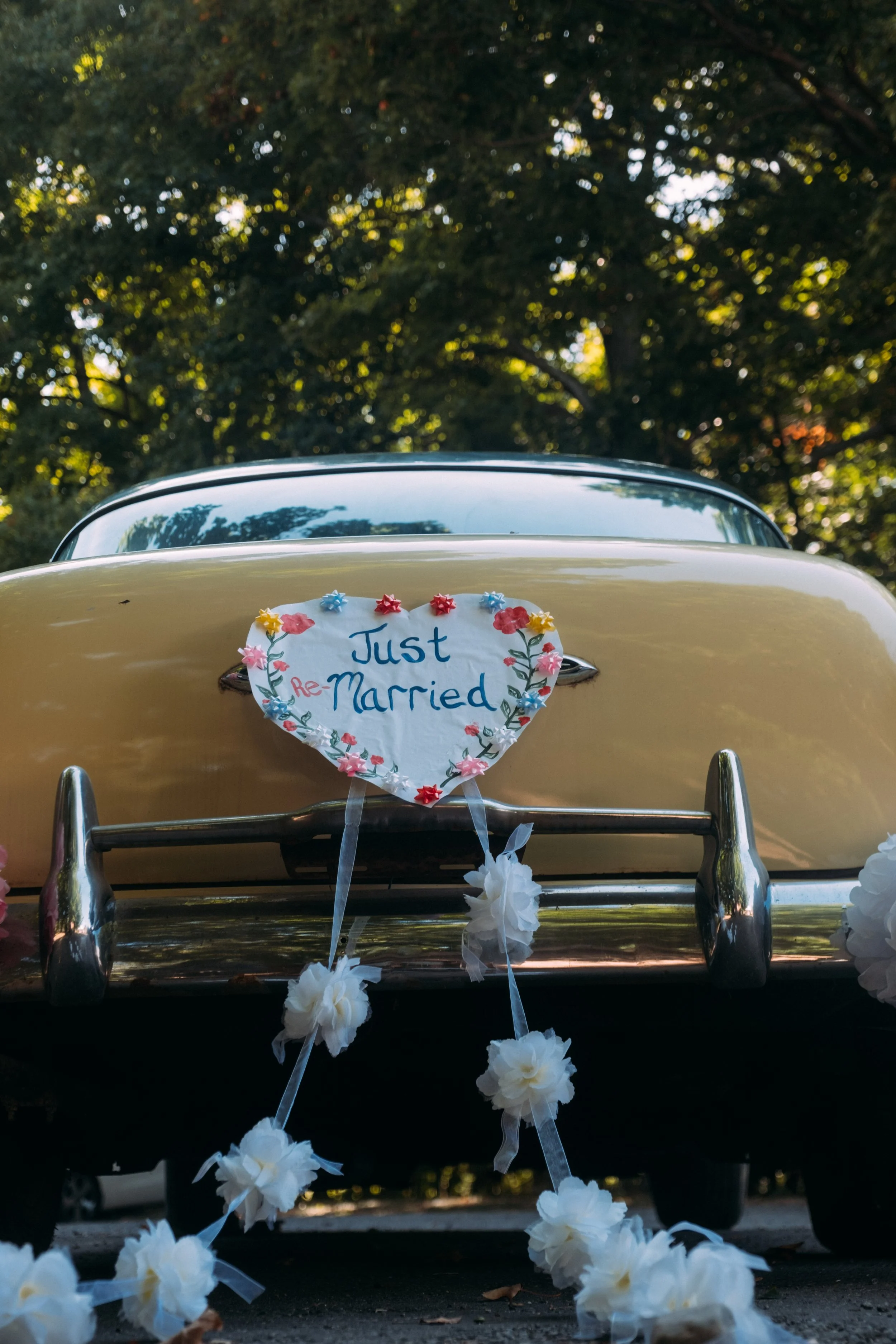 Octagonal yellow vintage car decorated with a white heart-shaped sign that reads "Just Re-Married" and white floral garlands, parked with a background of trees.