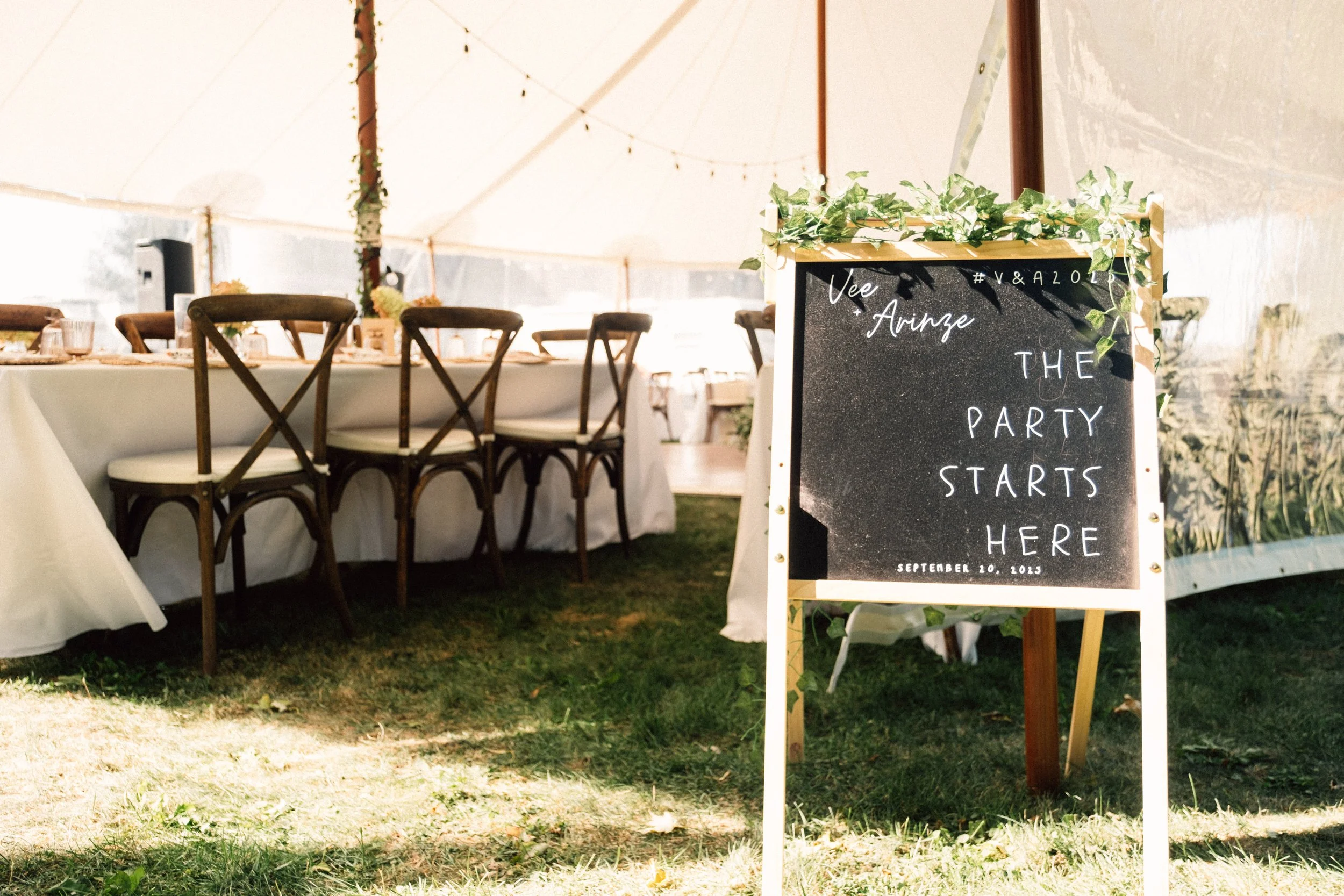 A black signboard with white handwritten text that reads 'The party starts here, September 20, 2023,' decorated with green leaves on top, standing outside under a tent near a long table with wooden chairs.