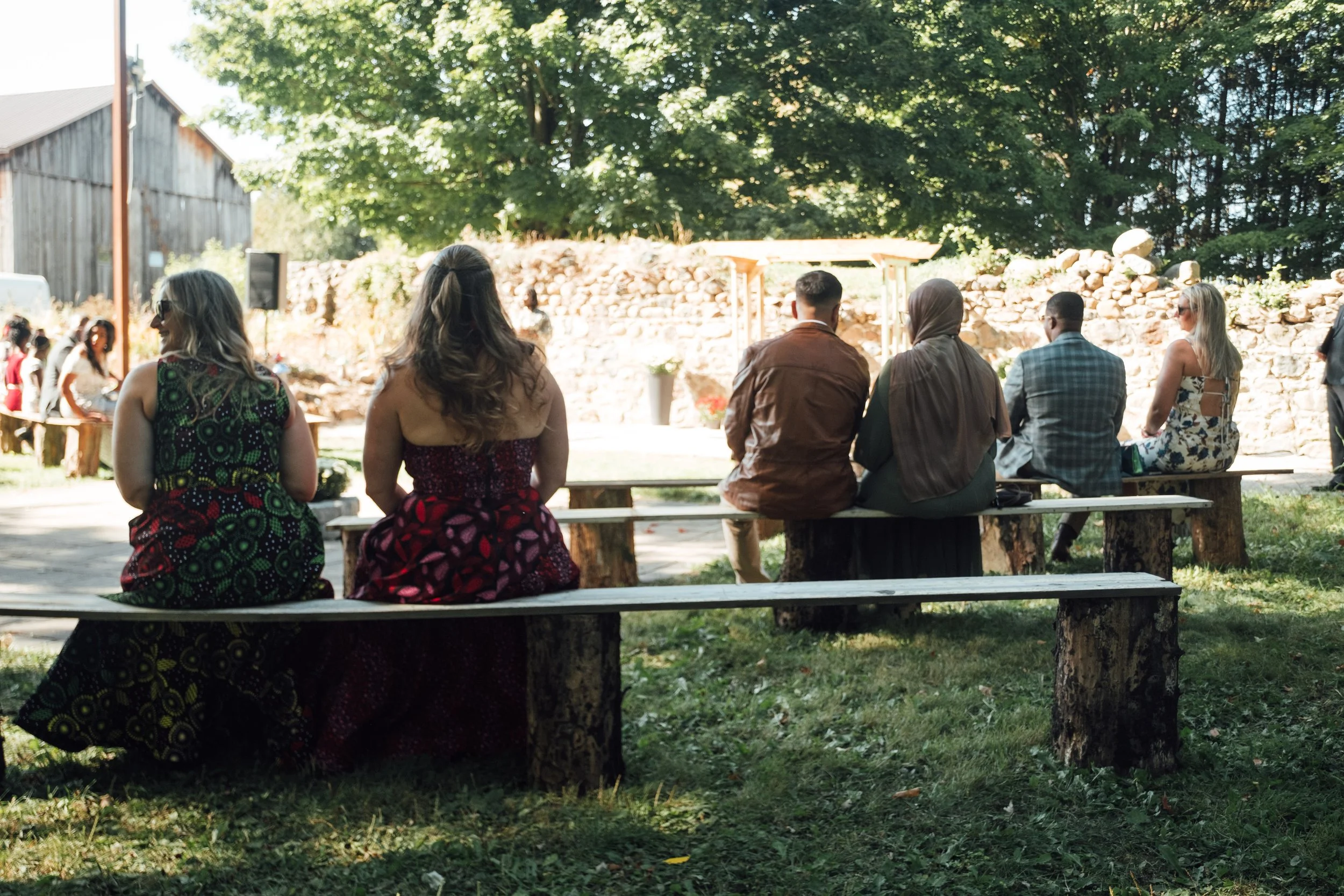 Group of people sitting on wooden benches outdoors, facing a stone wall and gathering in a park-like setting.