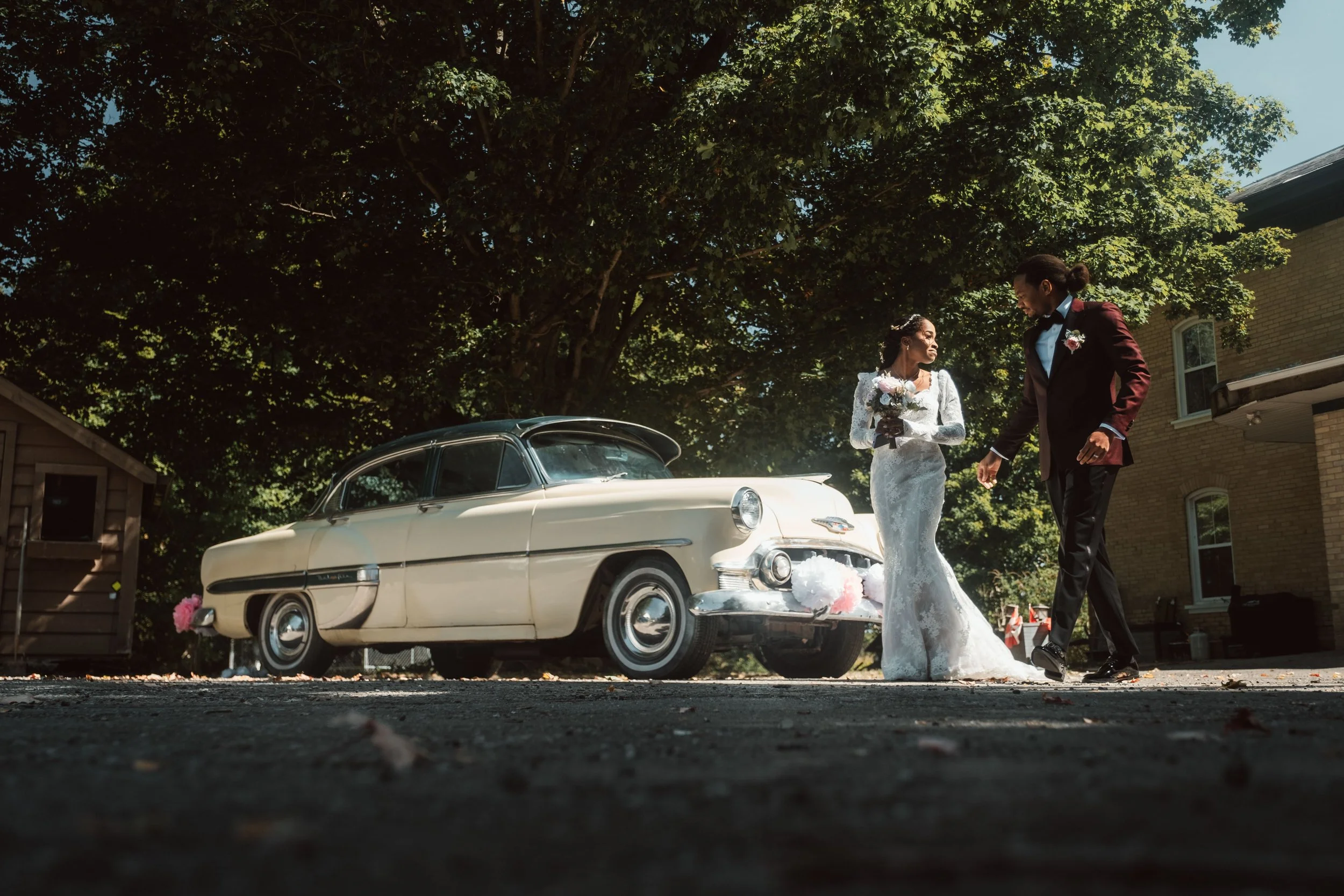 A bride and groom standing outdoors in front of a vintage cream-colored car, surrounded by green trees under blue sky, on a sunny day.