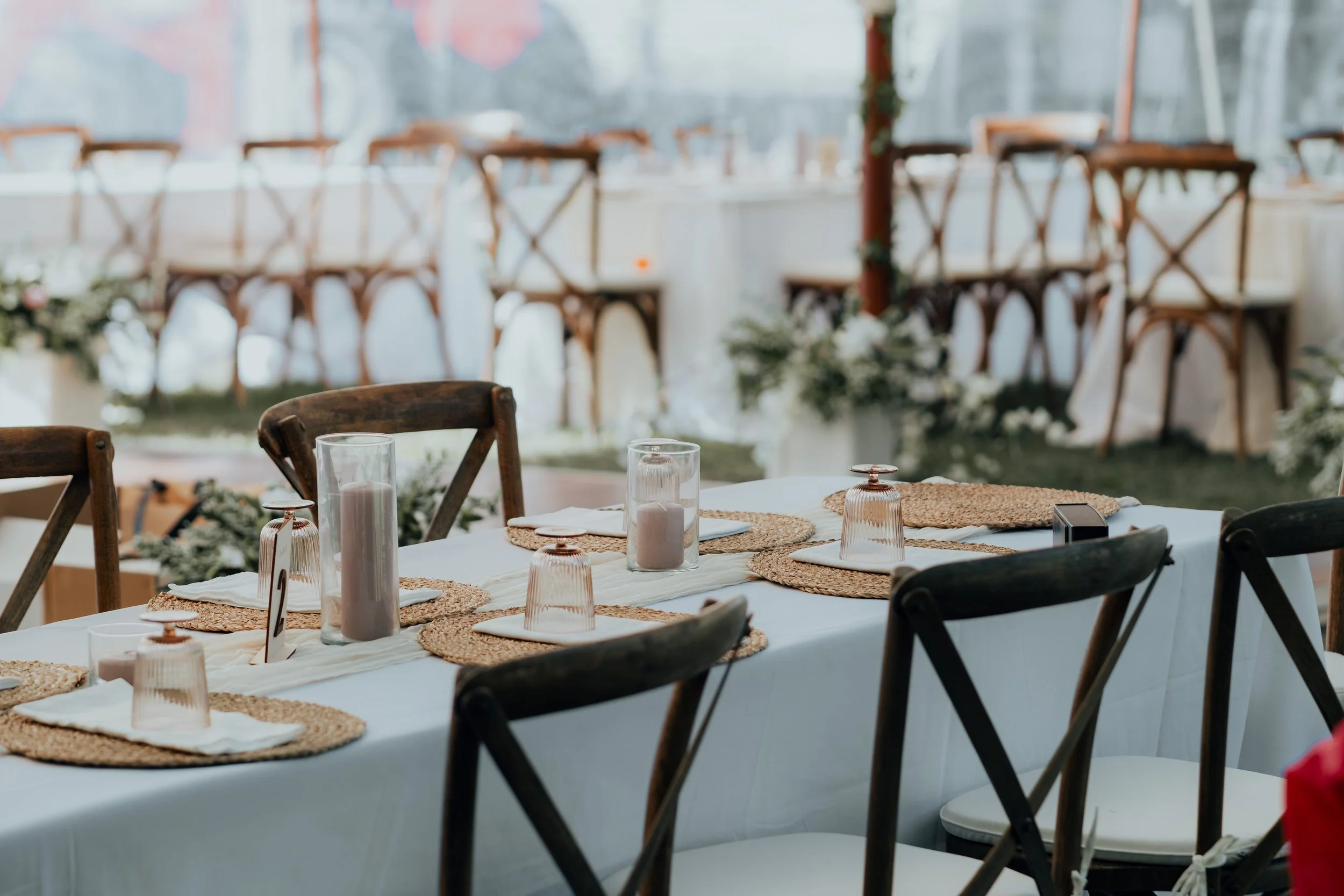 Decorated banquet table with white tablecloth, woven placemats, glass candleholders with candles, napkins, and table number, set for a wedding or celebration, with chairs and floral arrangements in the background.