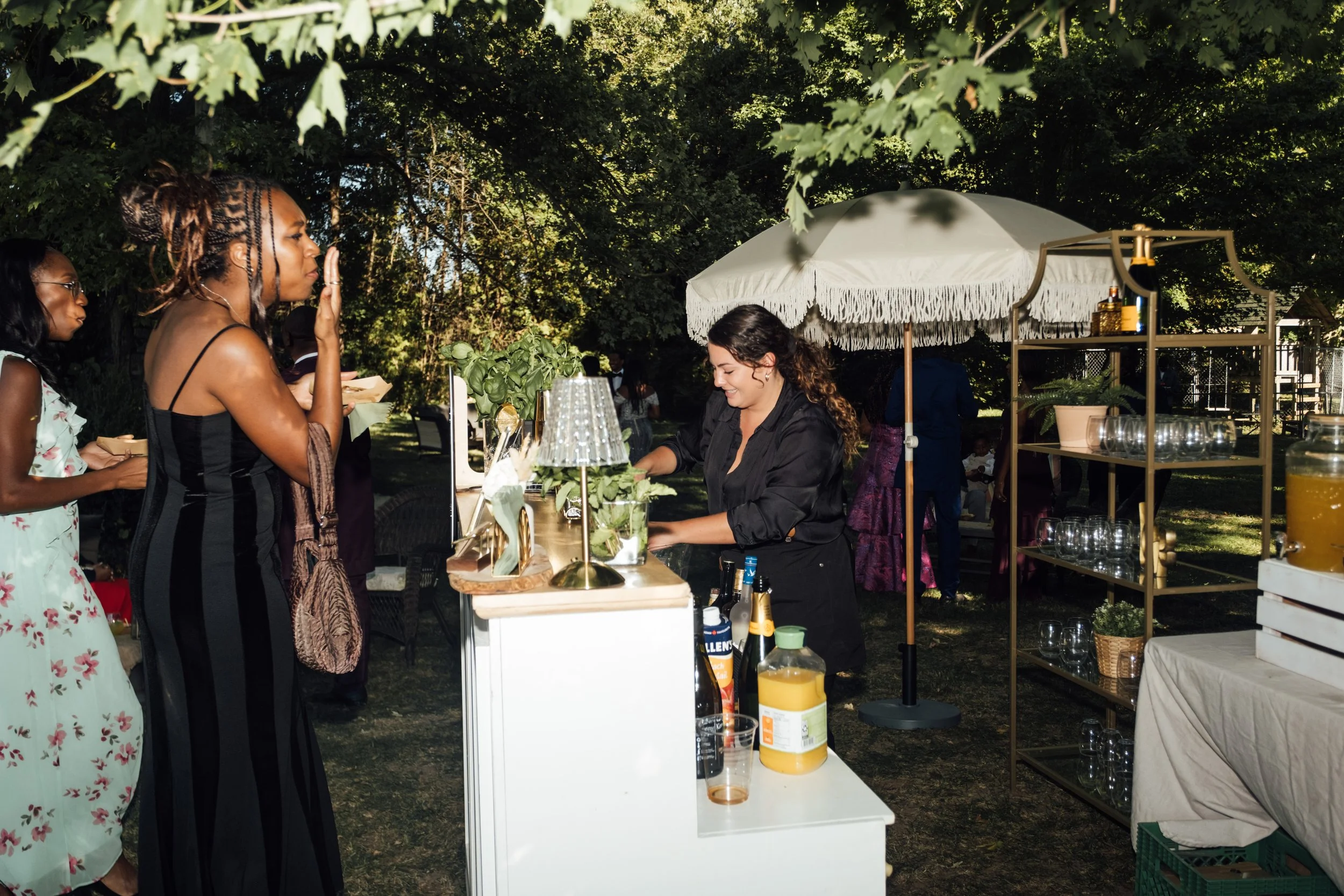 Women standing in line at an outdoor beverage stand, being served by a woman behind the counter under a large white umbrella, with green trees in the background.