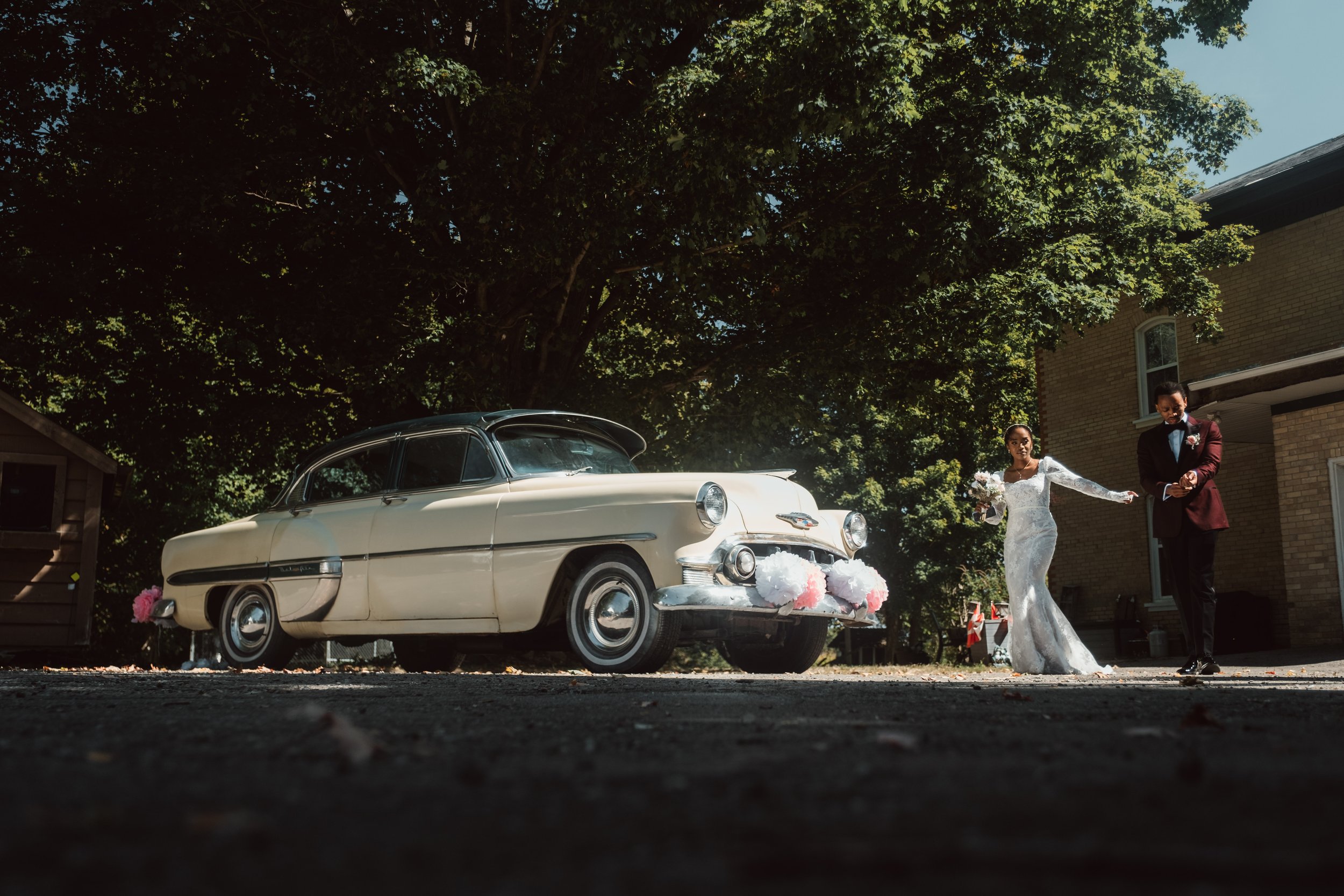 A bride and groom walking away from a vintage car decorated with pink and white flowers, outdoors under a large tree with a building in the background on a sunny day.