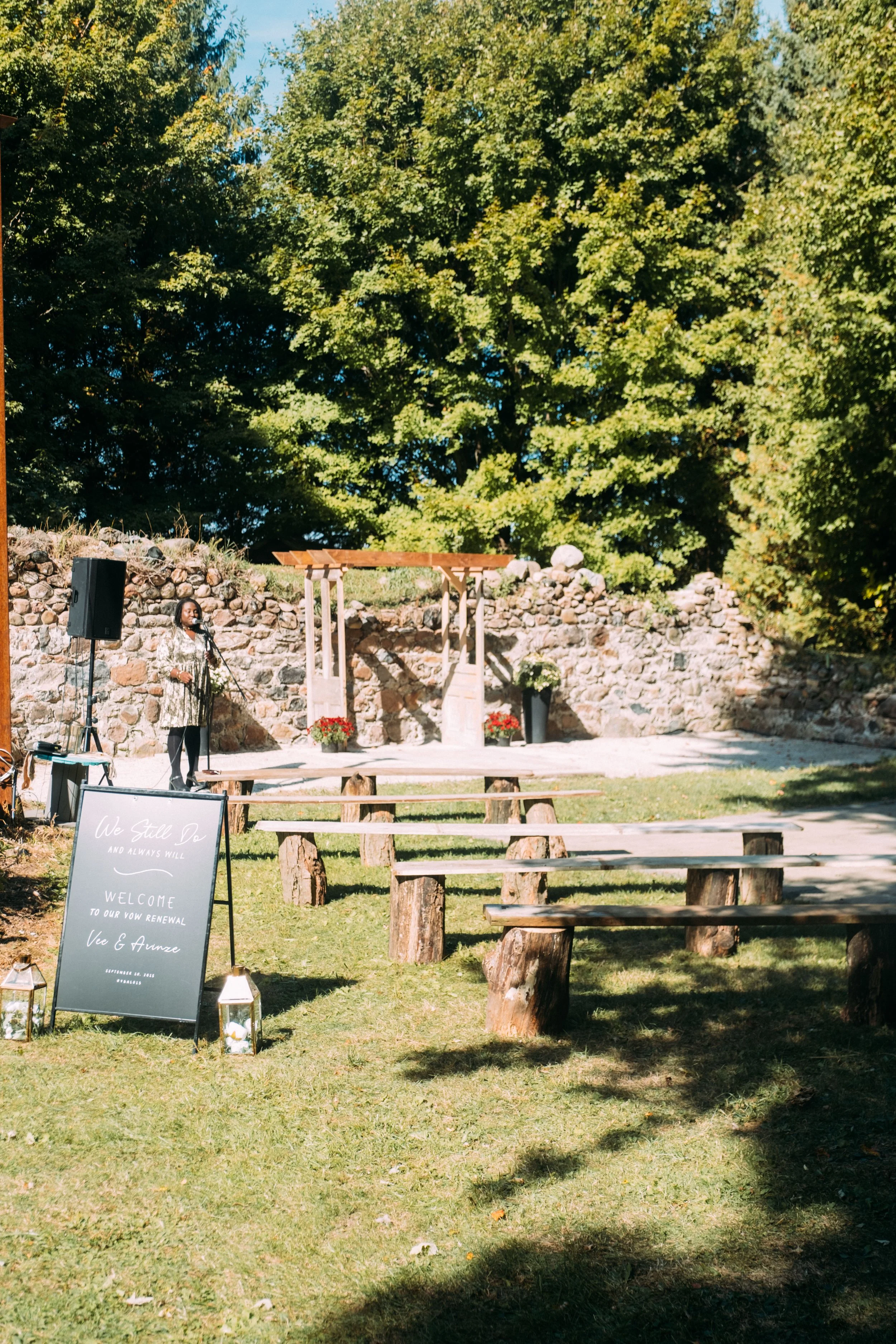 An outdoor wedding ceremony setup with wooden benches on a grassy area, a stone wall backdrop, decorated with flowers, and a woman speaking at a microphone near a welcome sign and lanterns.