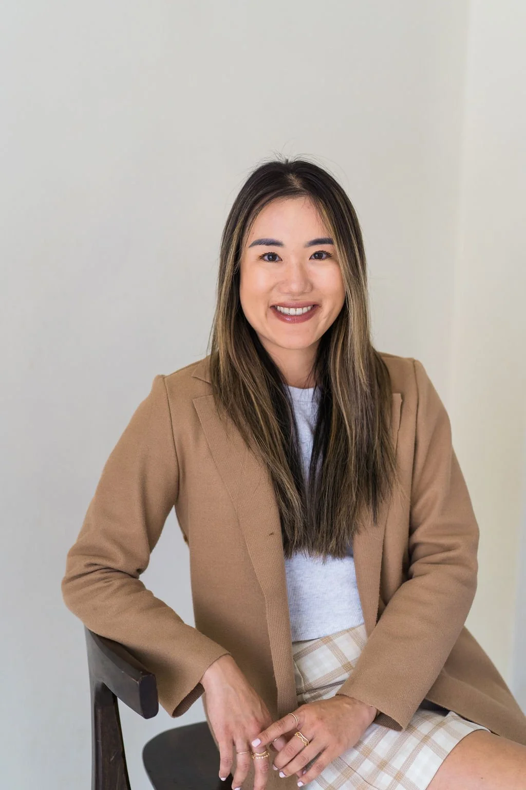 A woman with long dark hair and a big smile, wearing a tan blazer, white shirt, and plaid skirt, sitting on a chair against a plain light-colored wall.