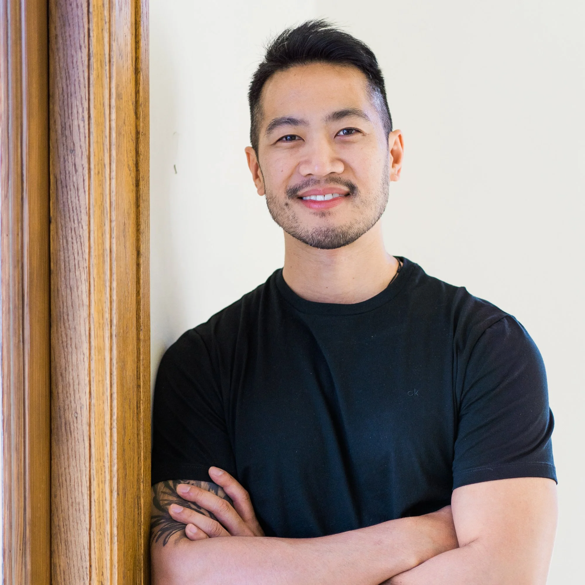 A smiling man with dark hair and a beard, wearing a black t-shirt, standing with arms crossed next to a wooden doorframe.
