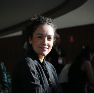 A young girl with curly hair smiling at the camera in a dimly lit indoor setting surrounded by other people.