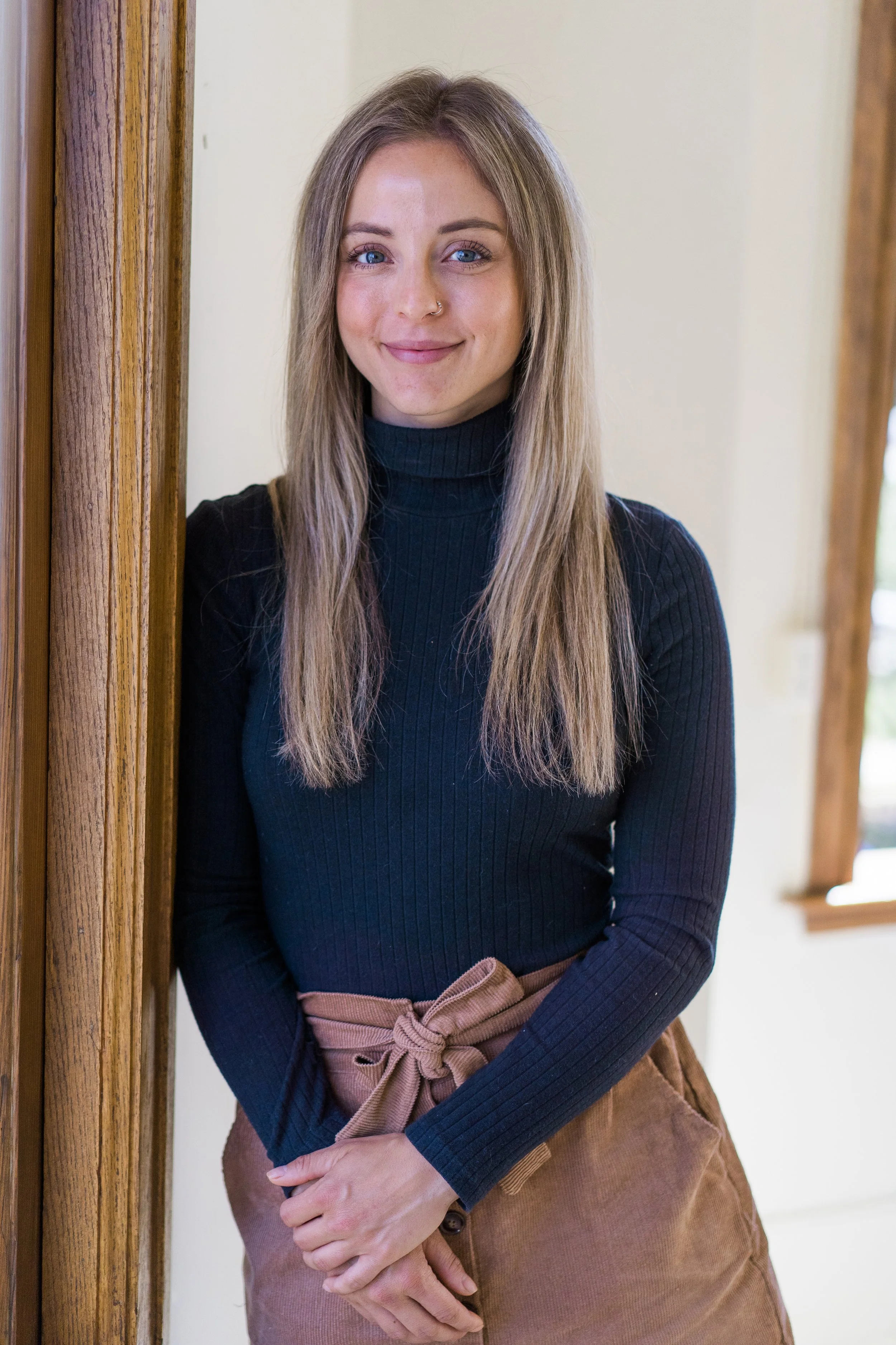 A young woman with long blonde hair, blue eyes, and a nose ring, standing indoors near a wooden doorframe, smiling at the camera.