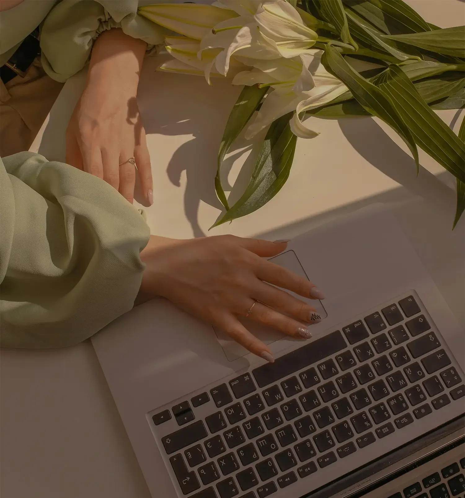 Person with manicured nails using a laptop keyboard, next to a bouquet of white lilies and green foliage on a white surface.