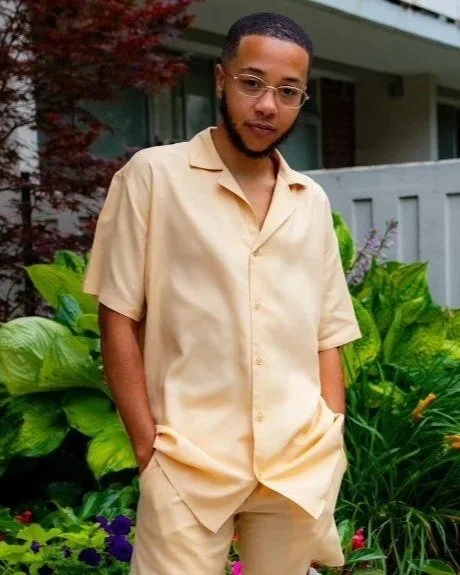 Young man with glasses and a beard standing outdoors near green plants, wearing a beige button-up shirt and beige pants.
