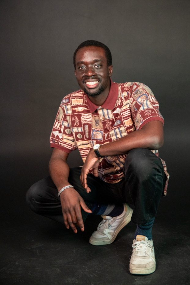 Man crouching, smiles, wearing patterned shirt, dark pants, white sneakers, dark background.