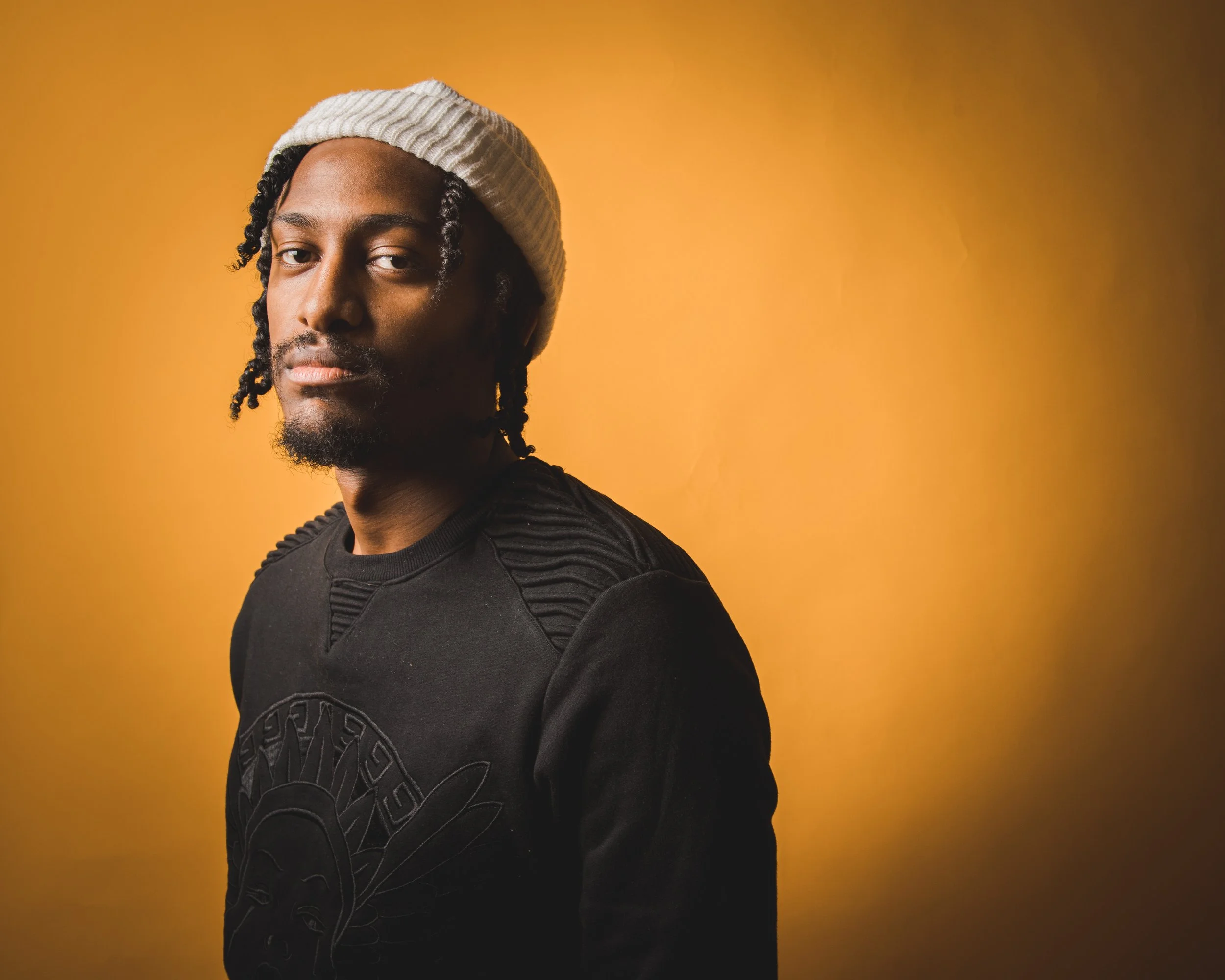 A man with dreadlocks, wearing a white knit beanie and a black sweatshirt with a graphic design, poses against a yellow background.