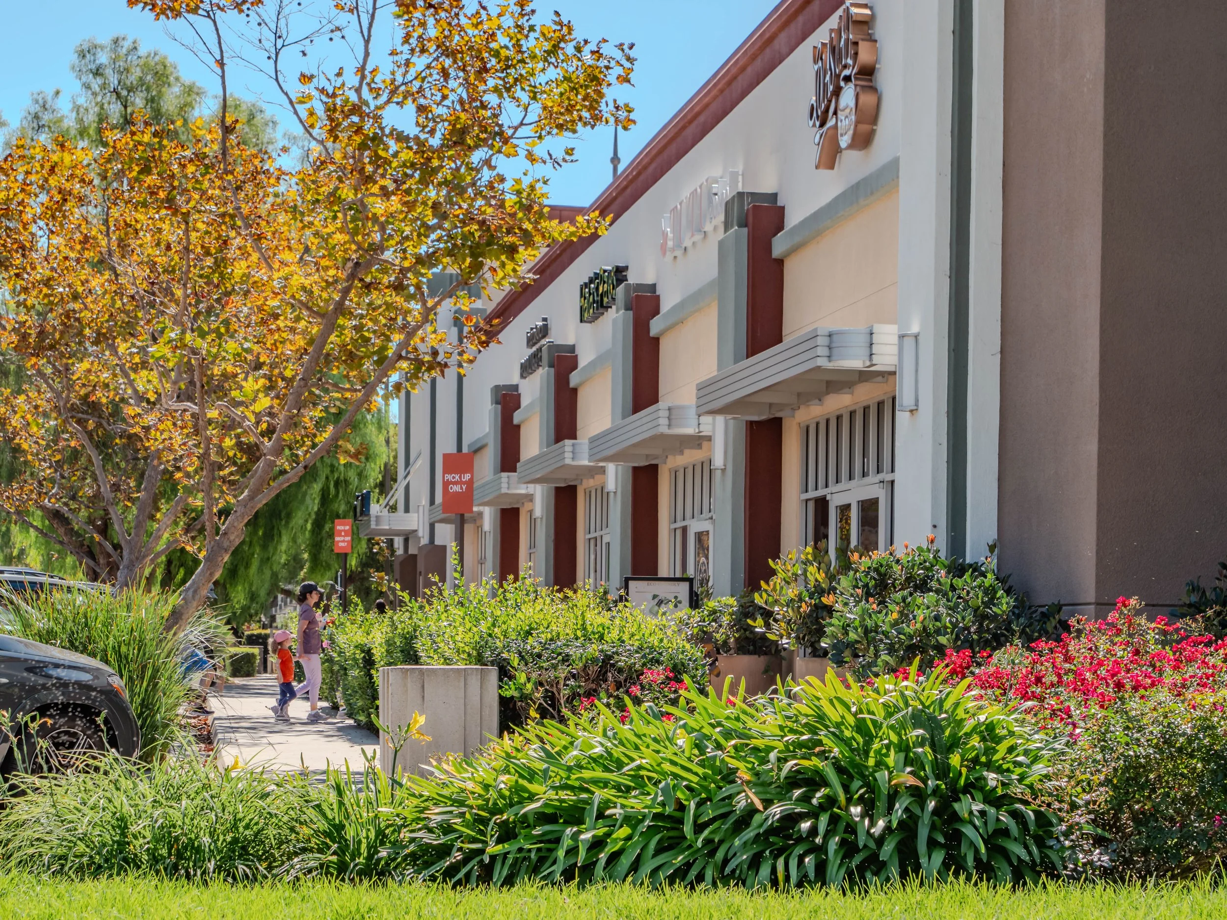 A shopping center with a row of stores, including Taco Bell and other fast-food restaurants, is seen under a blue sky. There are trees with autumn-colored leaves and landscaped bushes and flowers in the foreground. Two people, possibly a woman and a 