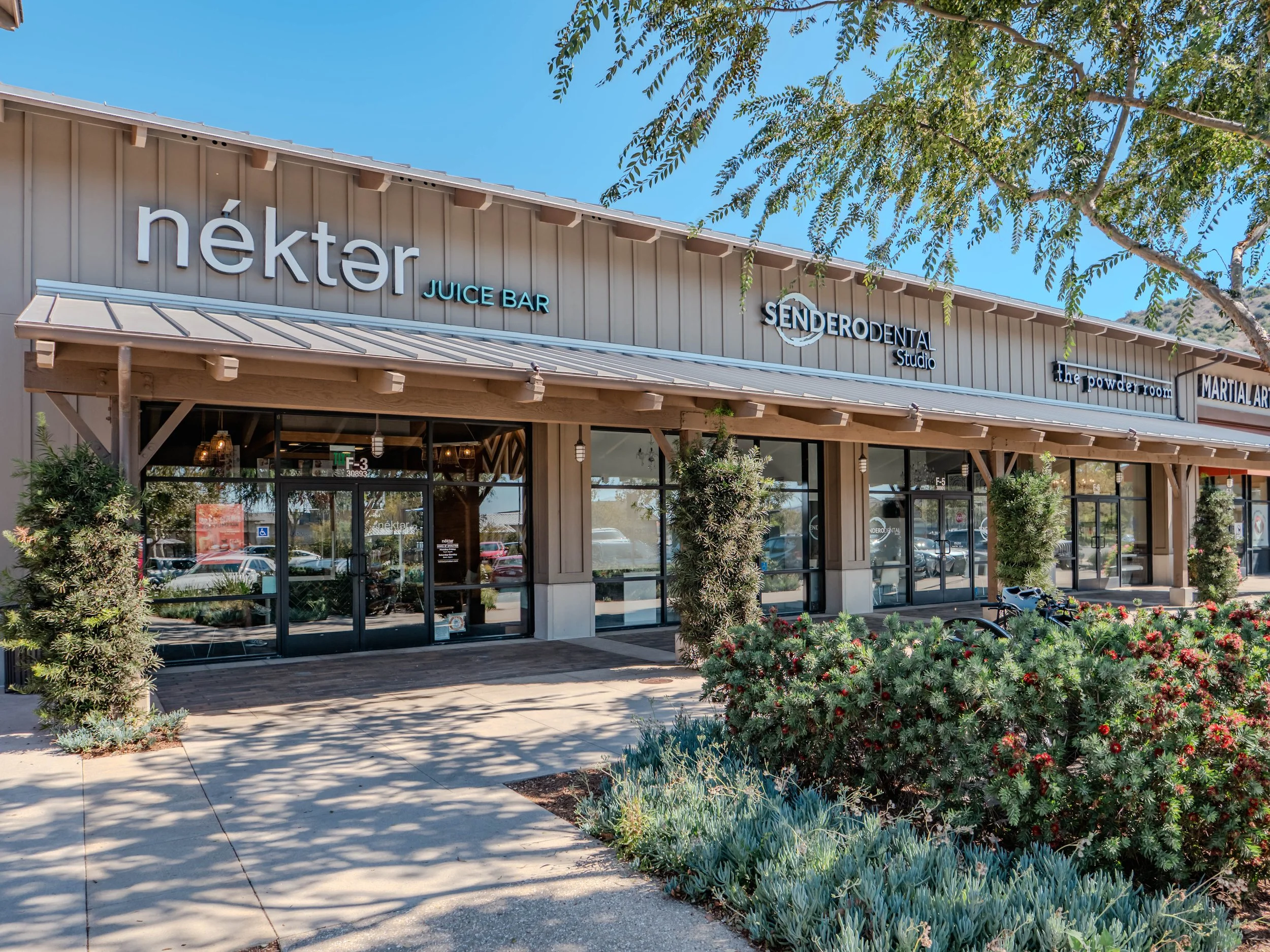 Exterior of a storefront shopping plaza with multiple business signs, including Nékter Juice Bar, Sender Odental Studio, The Powder Room, and Martial Arts, framed by trees and plants on a sunny day.