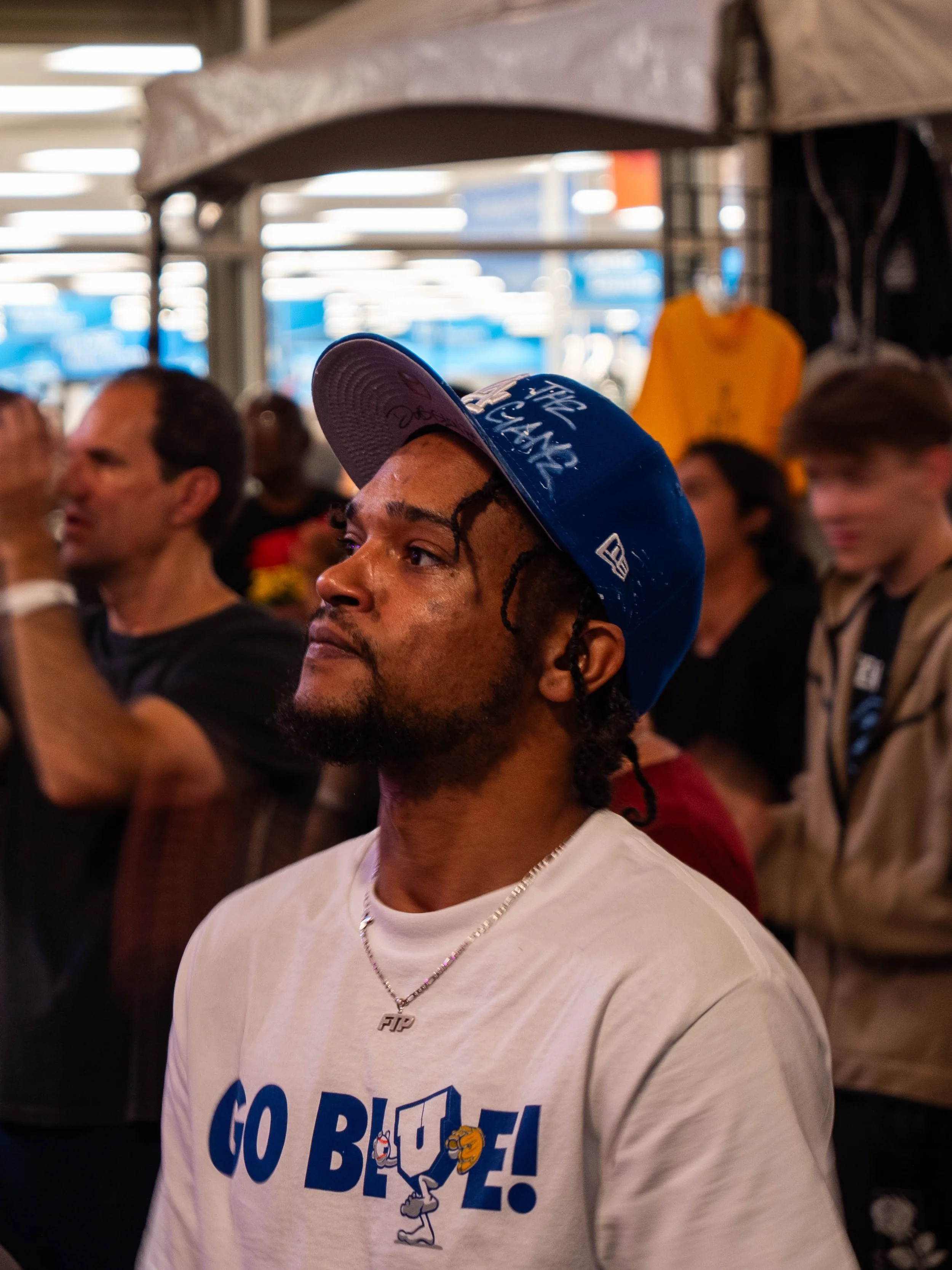 A man wearing a blue cap and white T-shirt with text and cartoon characters, standing in a crowd indoors with some people in the background.
