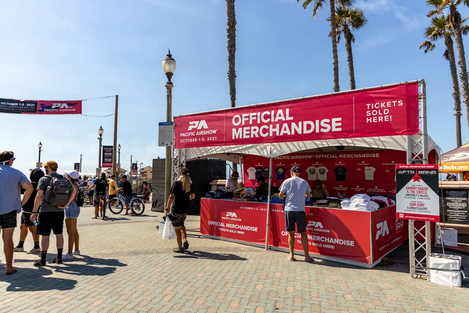 People at a merchandise booth with red and white signage reading 'Pacific Airshow Official Merchandise', selling T-shirts and other items, outdoors on a sunny day with palm trees in the background.