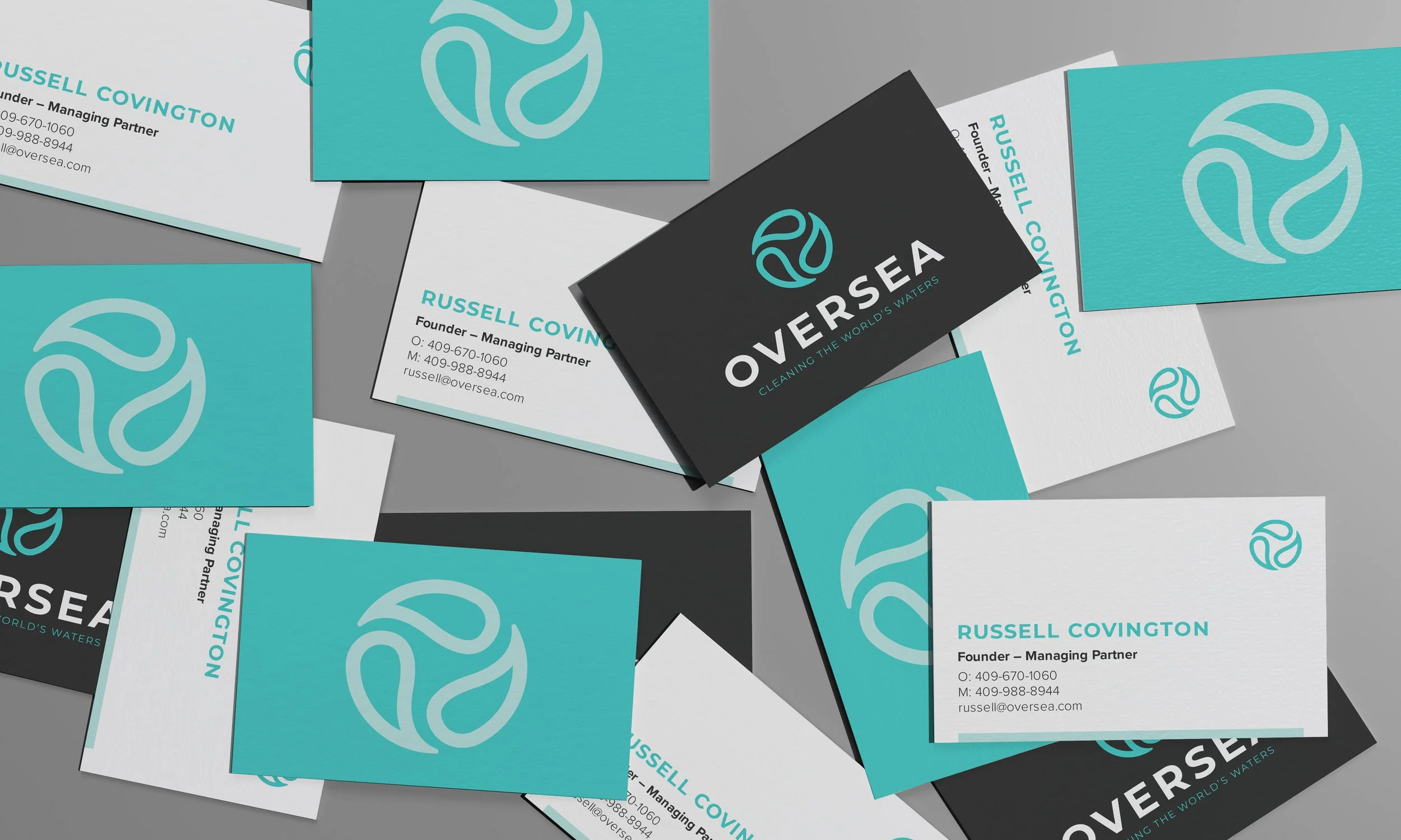 Business cards scattered on a gray surface featuring a logo with flowing lines and the text 'OVERSEA' and ‘CLEANING THE WORLD'S WATERS.’ The cards are in teal, black, and white with contact information for Russell Covington.