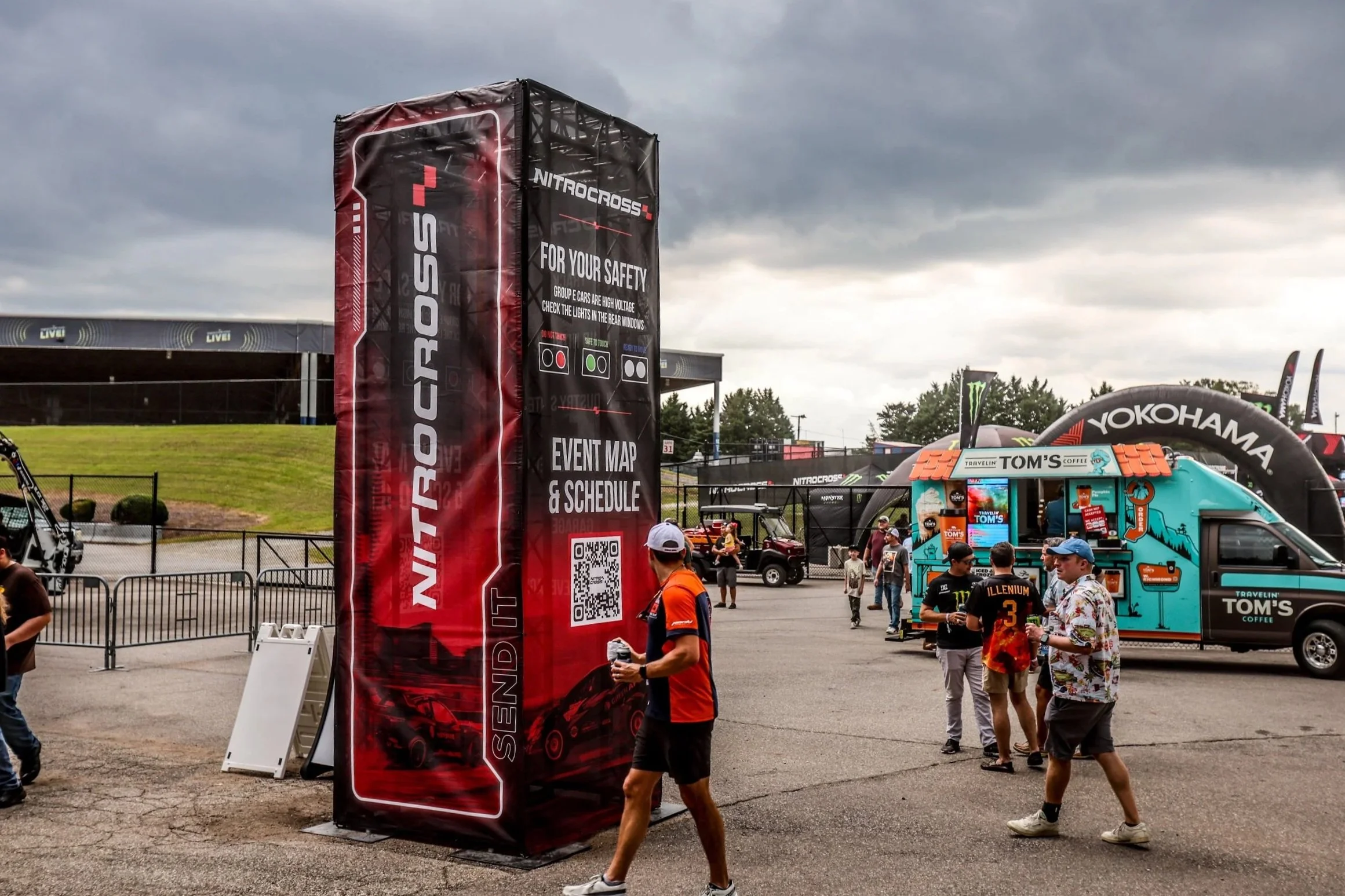 At an outdoor motorsport event, a large red and black NitroCross banner provides event information, QR code, and safety checks. Several attendees walk and chat near a colorful coffee truck and other event structures under a cloudy sky.