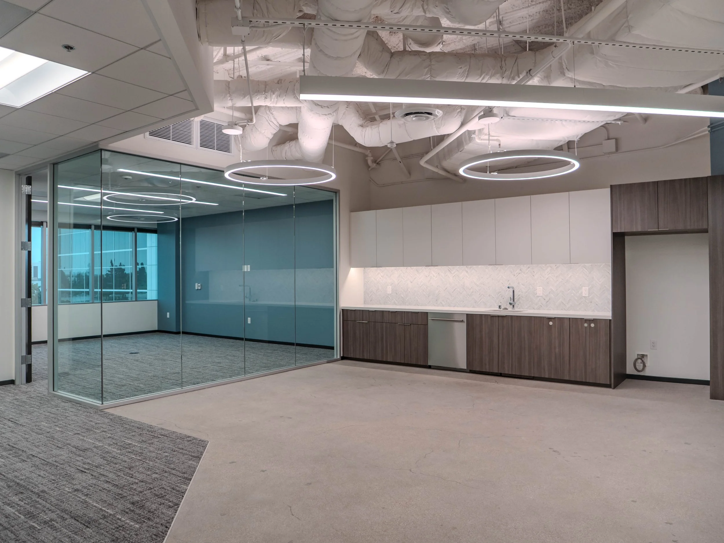 Empty office kitchen area with cabinets, countertop, and sink, separated by glass walls, with modern circular ceiling lights and exposed ceiling pipes.