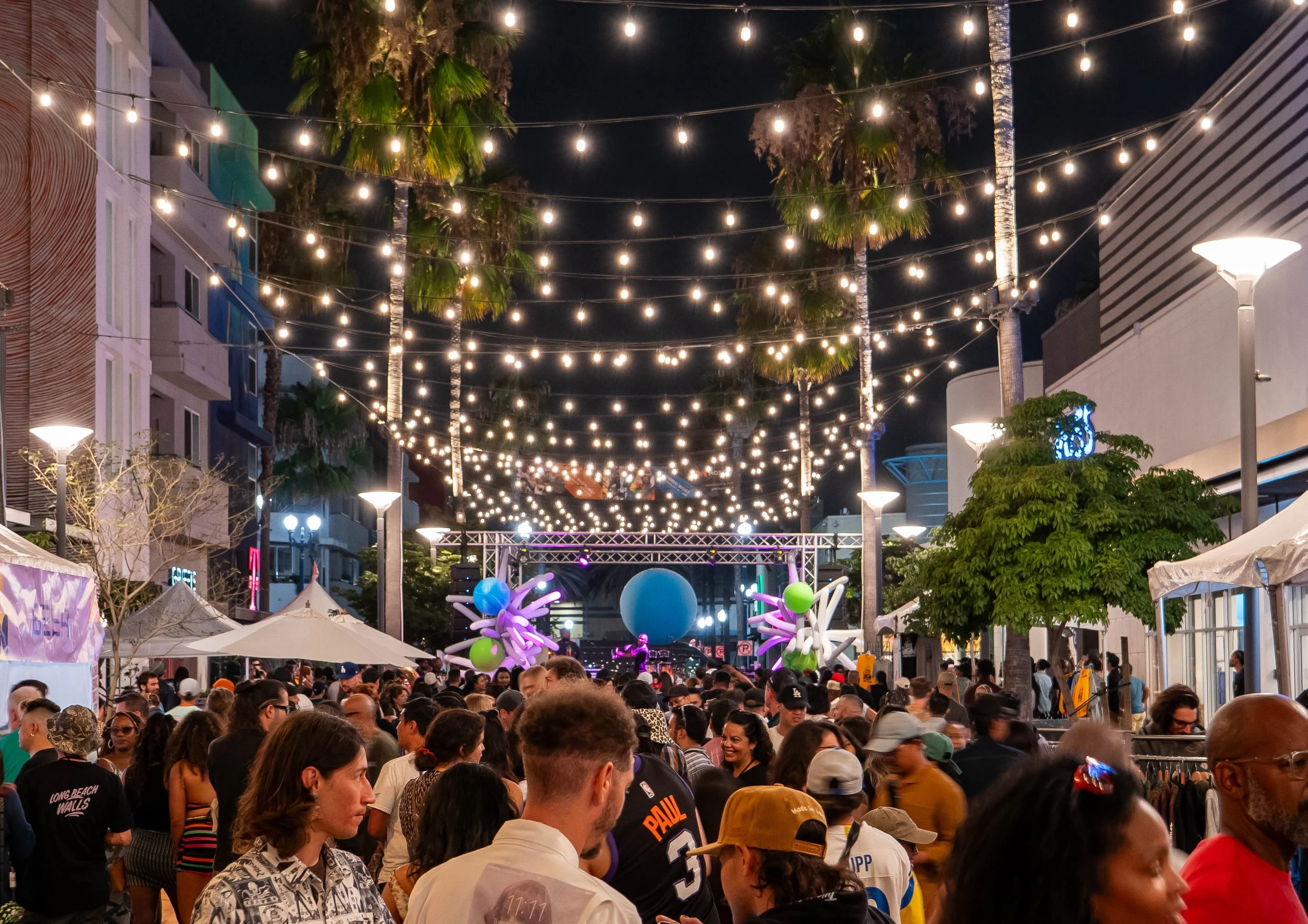 Outdoor night scene at a lively festival with a large crowd of people, string lights overhead, palm trees, and colorful balloon sculptures near a stage.