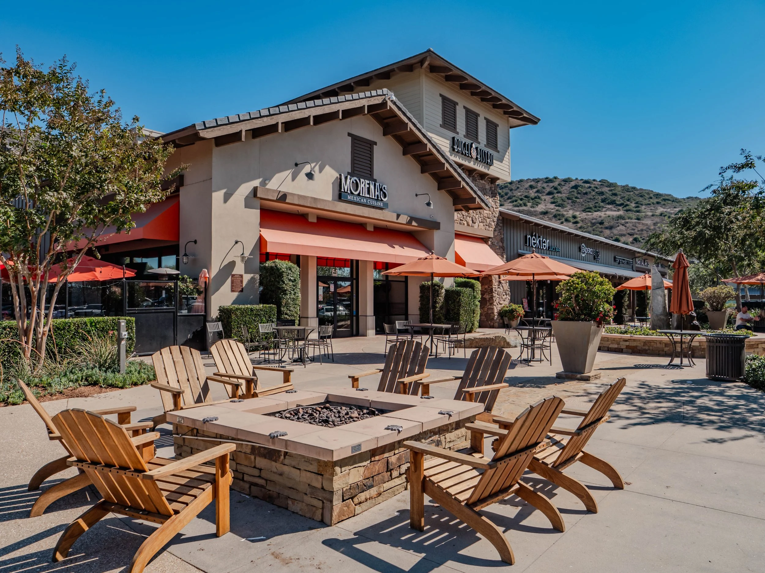 Outdoor seating area with wooden chairs surrounding a fire pit in front of shops with red awnings.