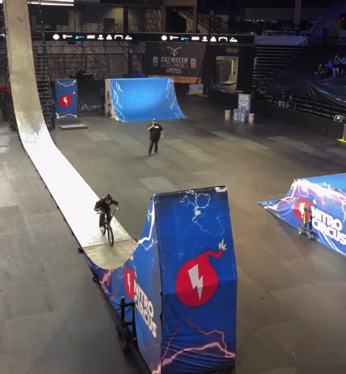 Indoor skate park with ramps and obstacles for skateboarding and bike tricks, featuring banners and displays with lightning and electrifying graphics, and a person riding a bike.