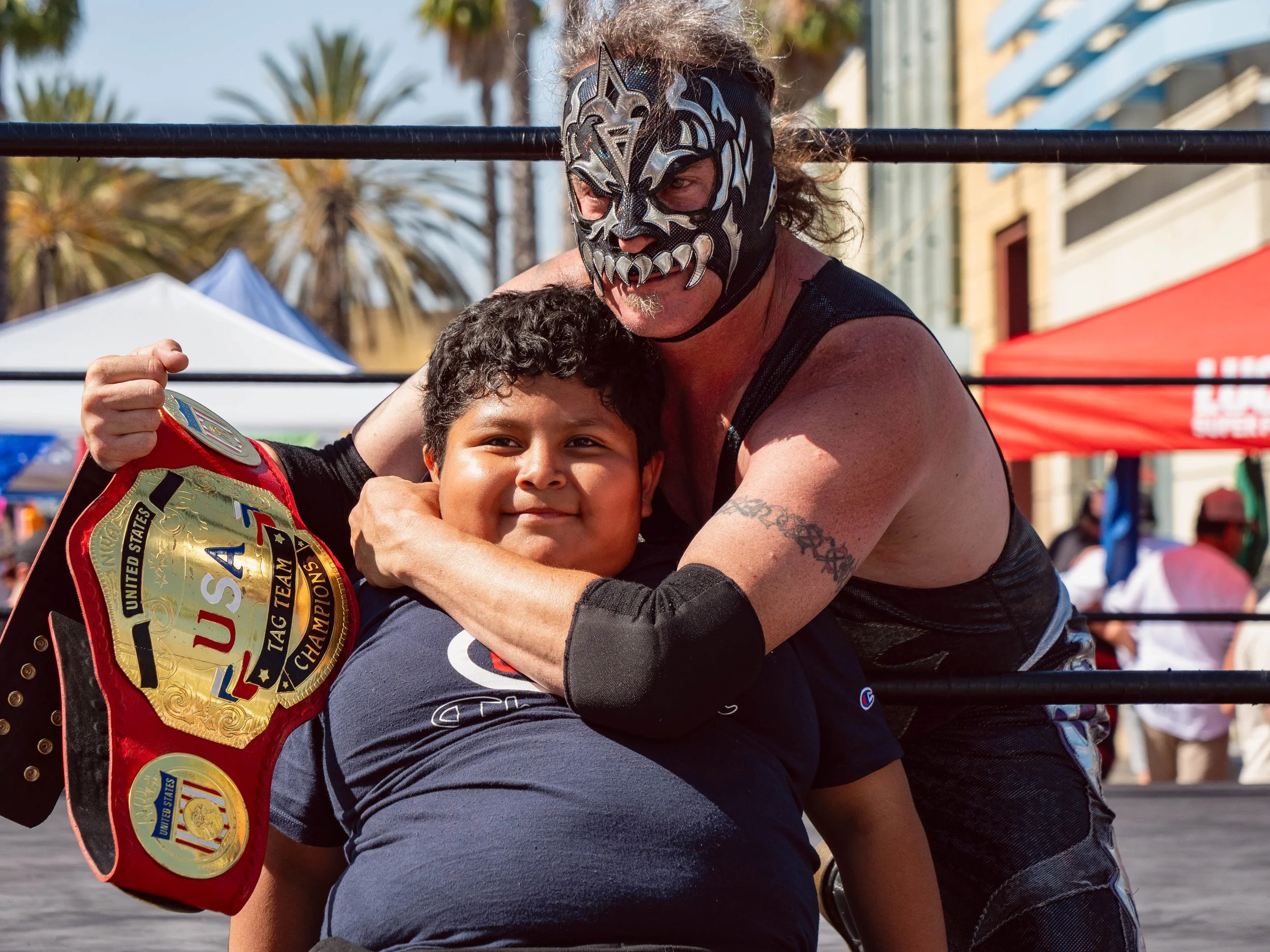 A wrestler with a jaguar mask hugging a young boy inside a wrestling ring. The boy is holding a United States champion belt. Crowd and tents are visible in the background.