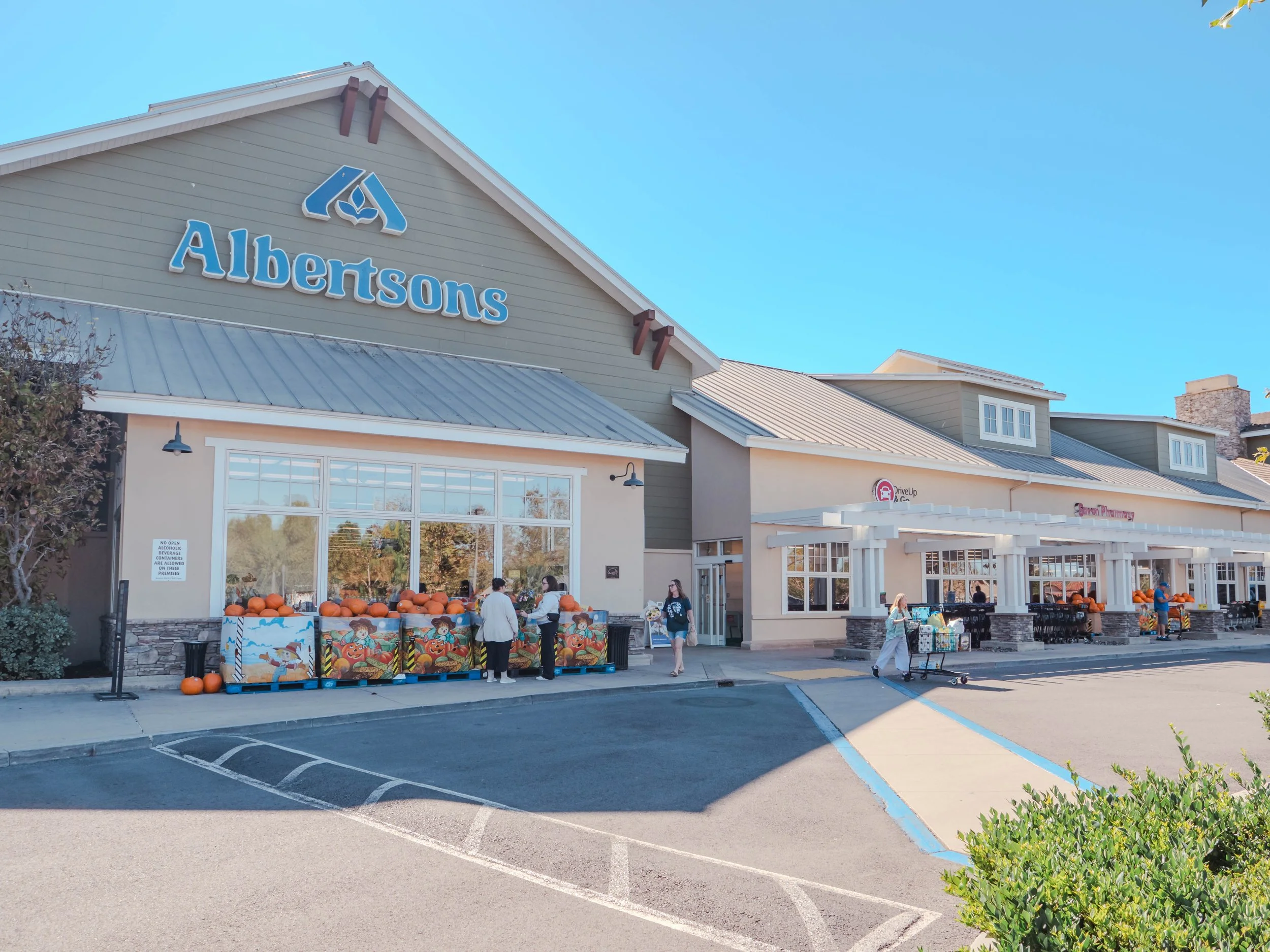 Exterior of an Albertsons grocery store with pumpkins displayed outside, customers shopping, and the store's sign visible on the building.
