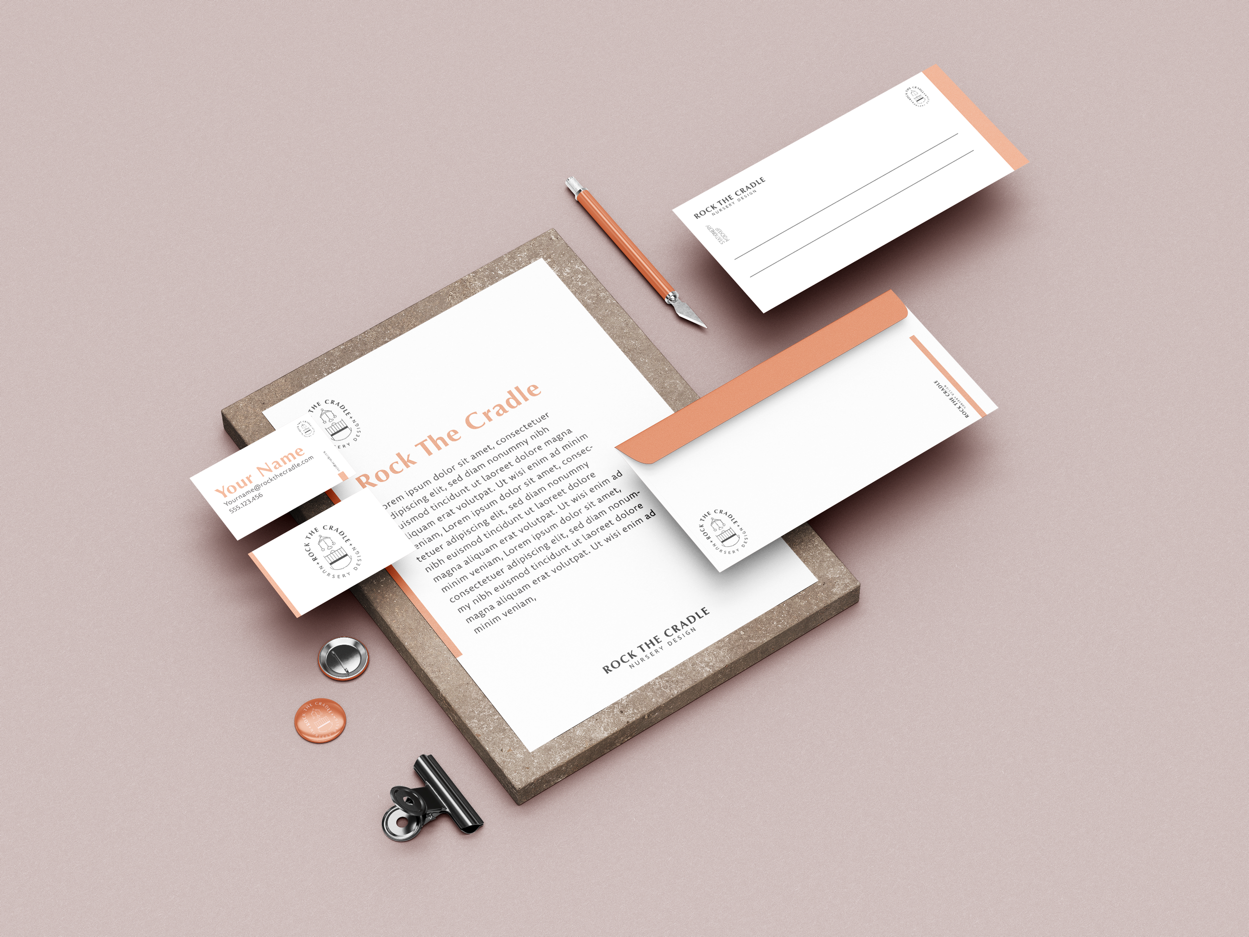 Set of branding materials for Rock The Cradle, including letterhead, business cards, and envelopes, in white and peach colors, arranged on a pink background with a pen, binder clip, and buttons nearby.