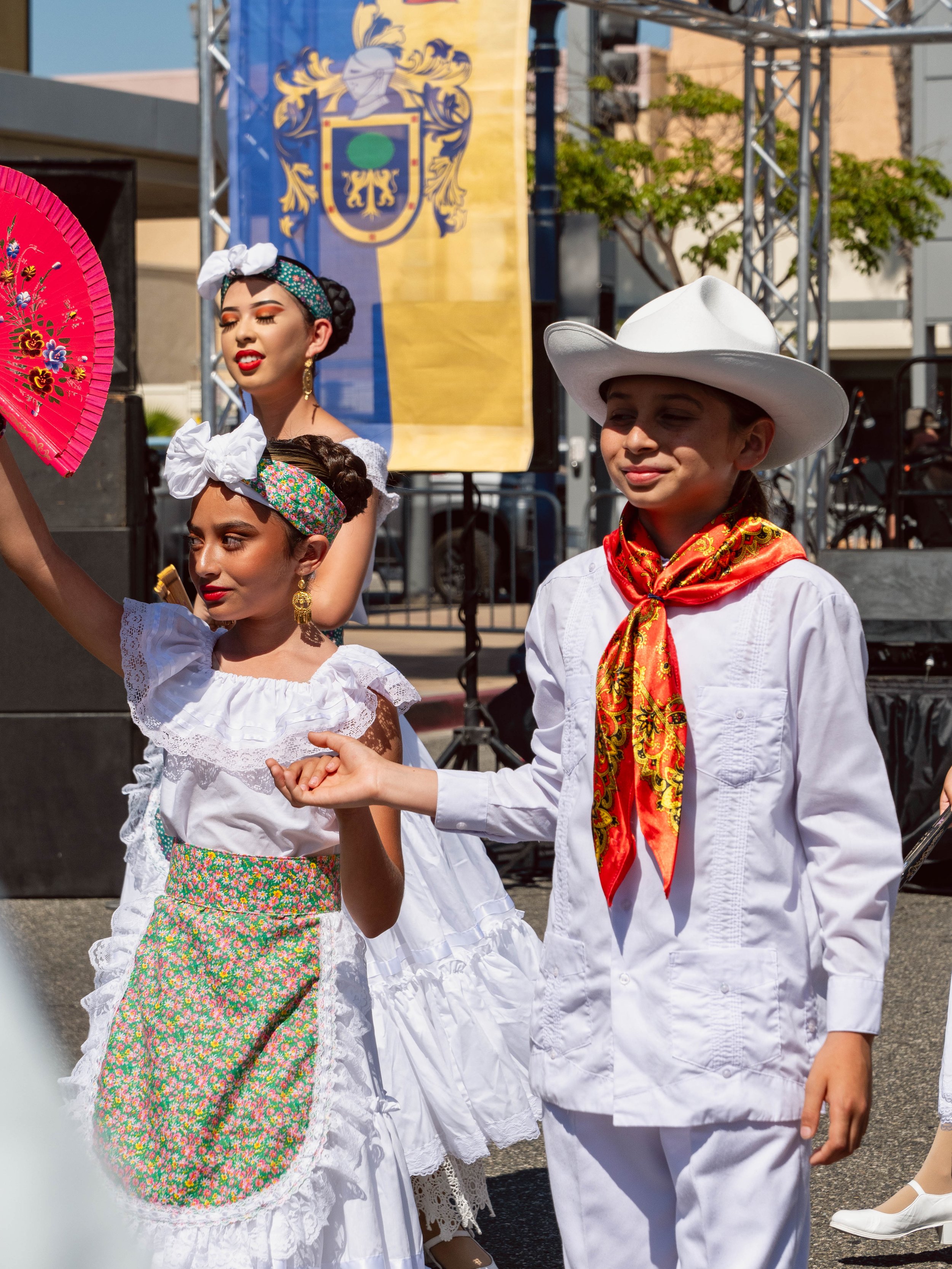 Children dressed in traditional Mexican attire participating in a cultural celebration outdoors. The girls wear white dresses with lace details, colorful headbands, and hold a pink fan. The boy wears a white cowboy hat, a white shirt, and a red banda