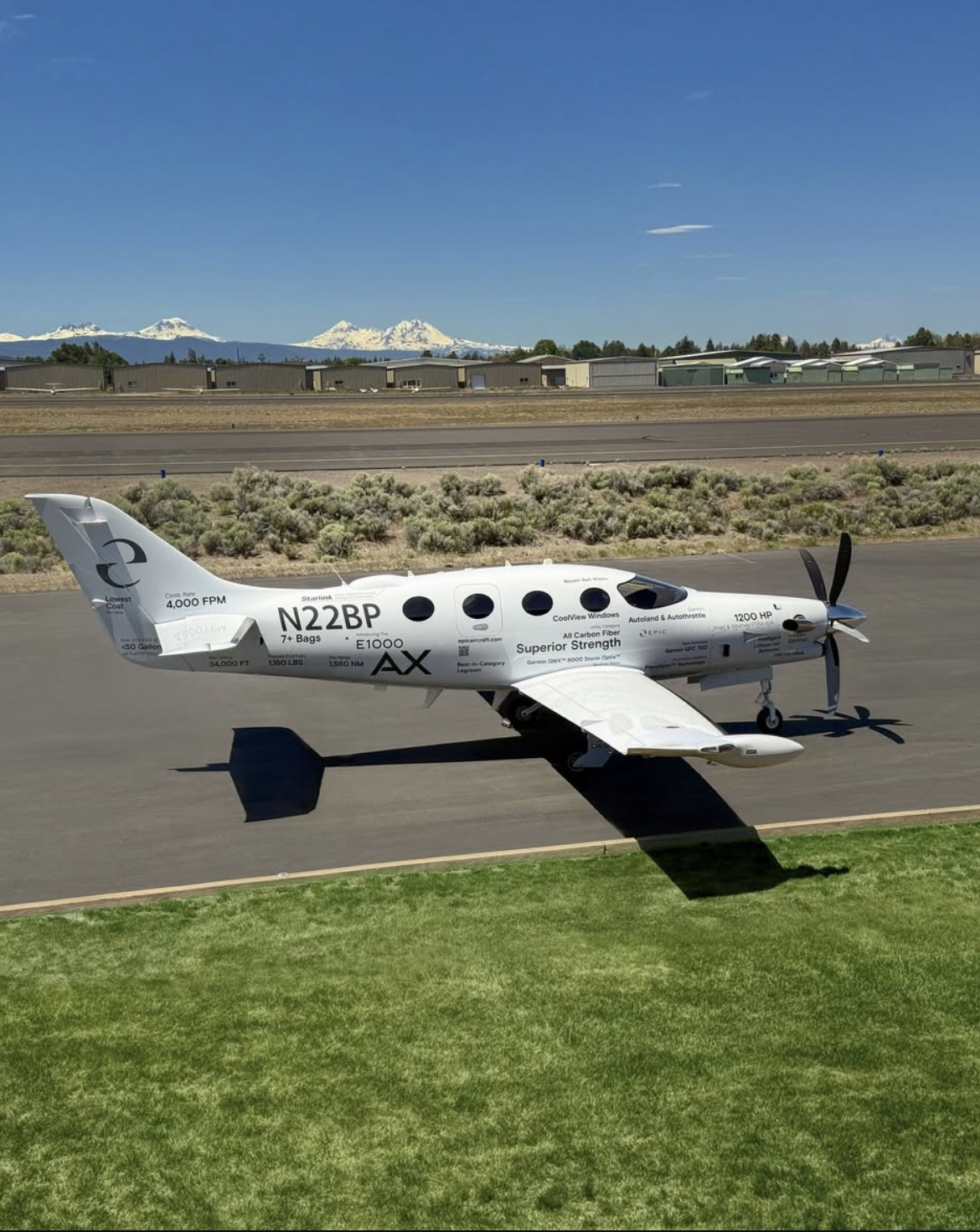 A white autonomous aerial drone with black text and logos on a tarmac, with a backdrop of distant snow-capped mountains and blue sky.