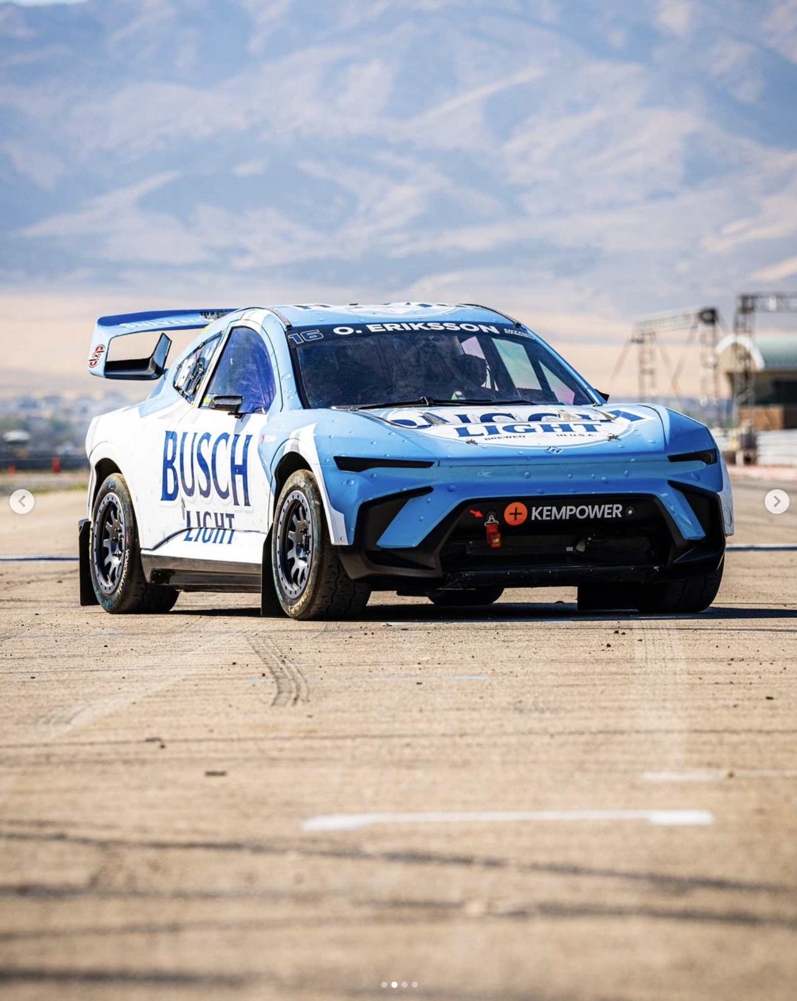 A race car with Volkswagen branding and Busch Light sponsorship, on a racetrack under a partly cloudy sky.