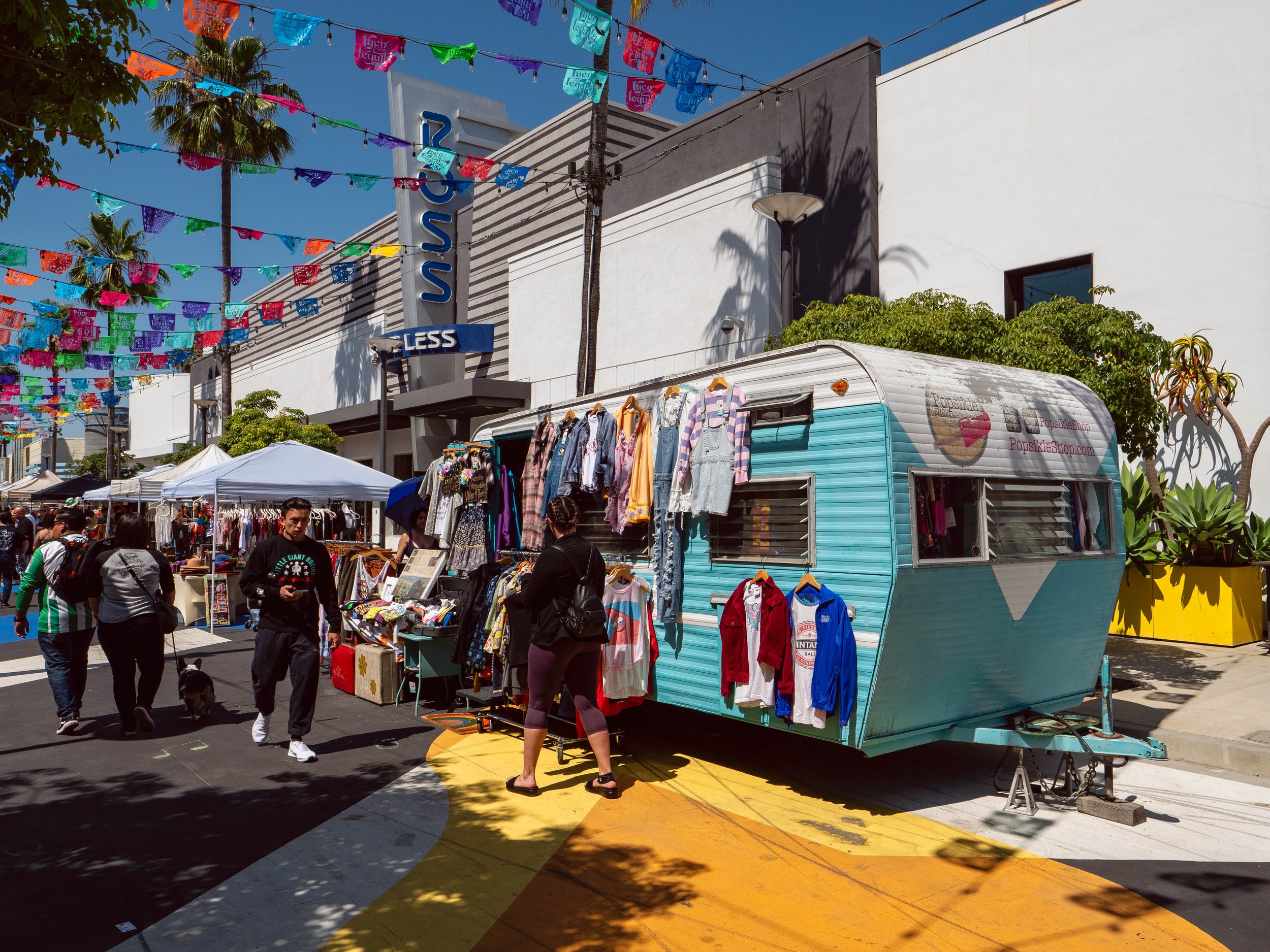 Street market with colorful flags overhead, small clothing stalls, and a vintage camper serving as a shop, in front of a modern shopping mall.