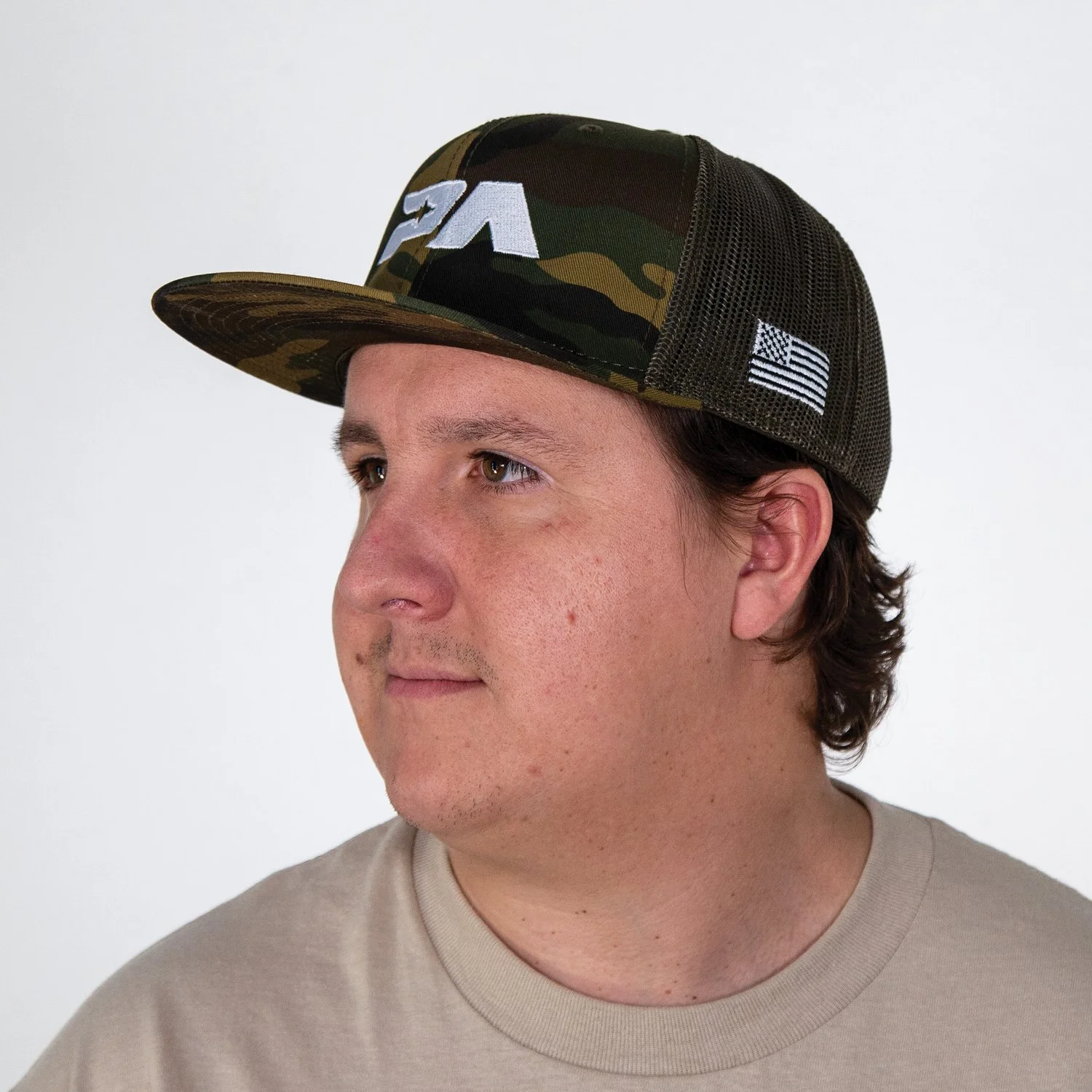 A young man wearing a camouflage baseball cap with a mesh back and American flag patch, looking to the side against a plain white background.
