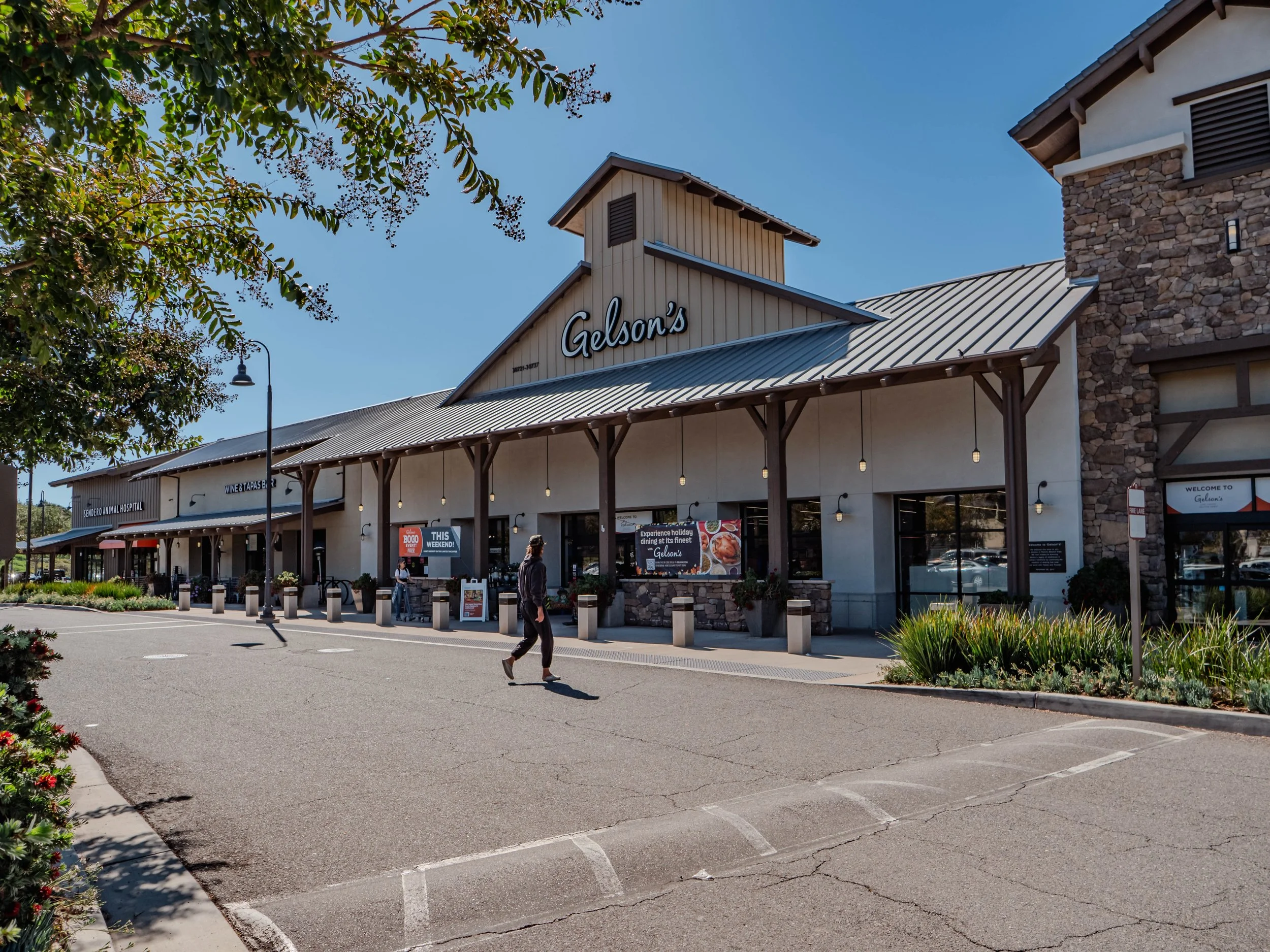 Outside view of Gelson's grocery store with parking lot, a few people walking, and clear blue sky.