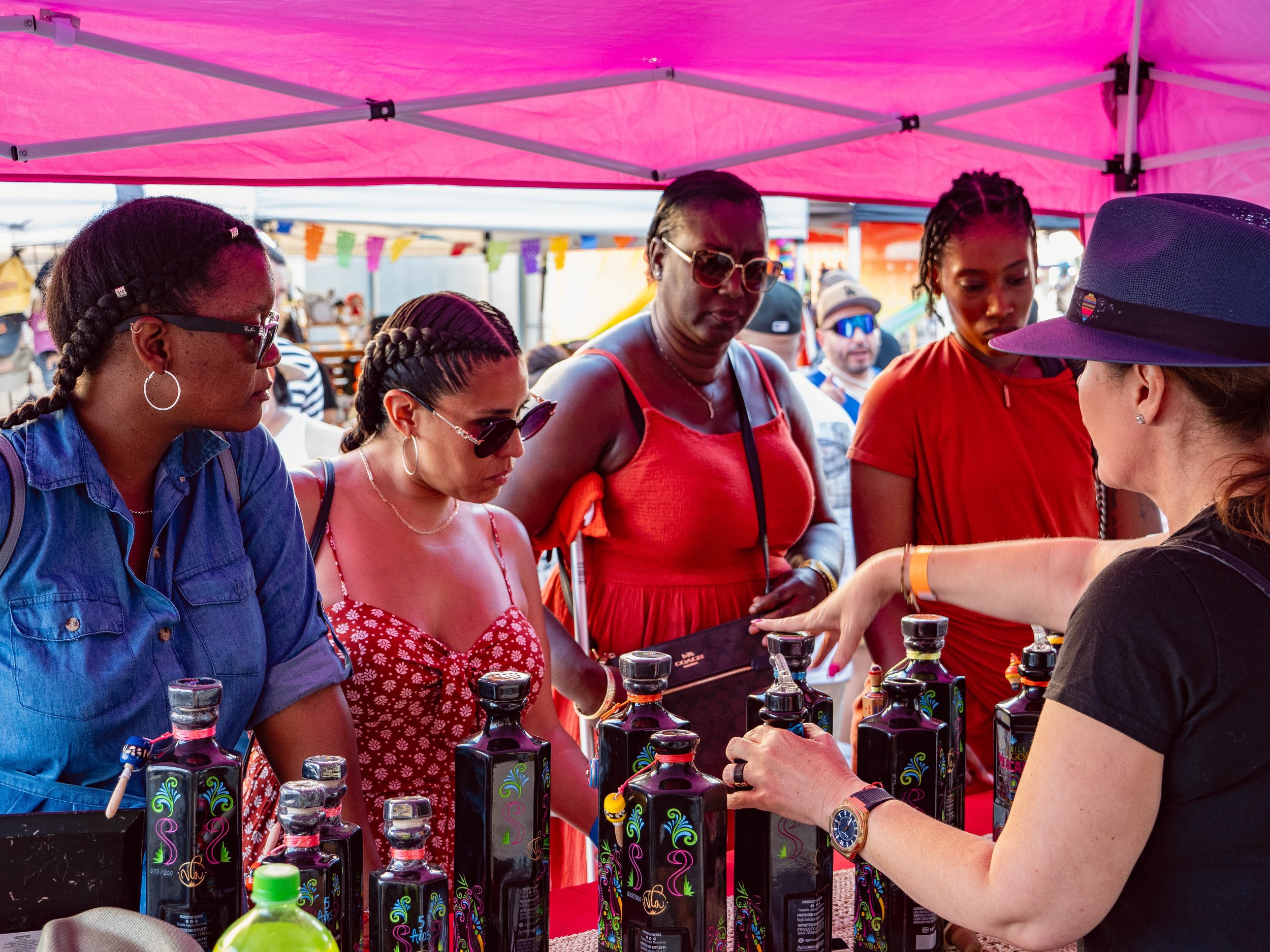 Group of women shopping for bottled liquor at an outdoor market stall with pink canopy.