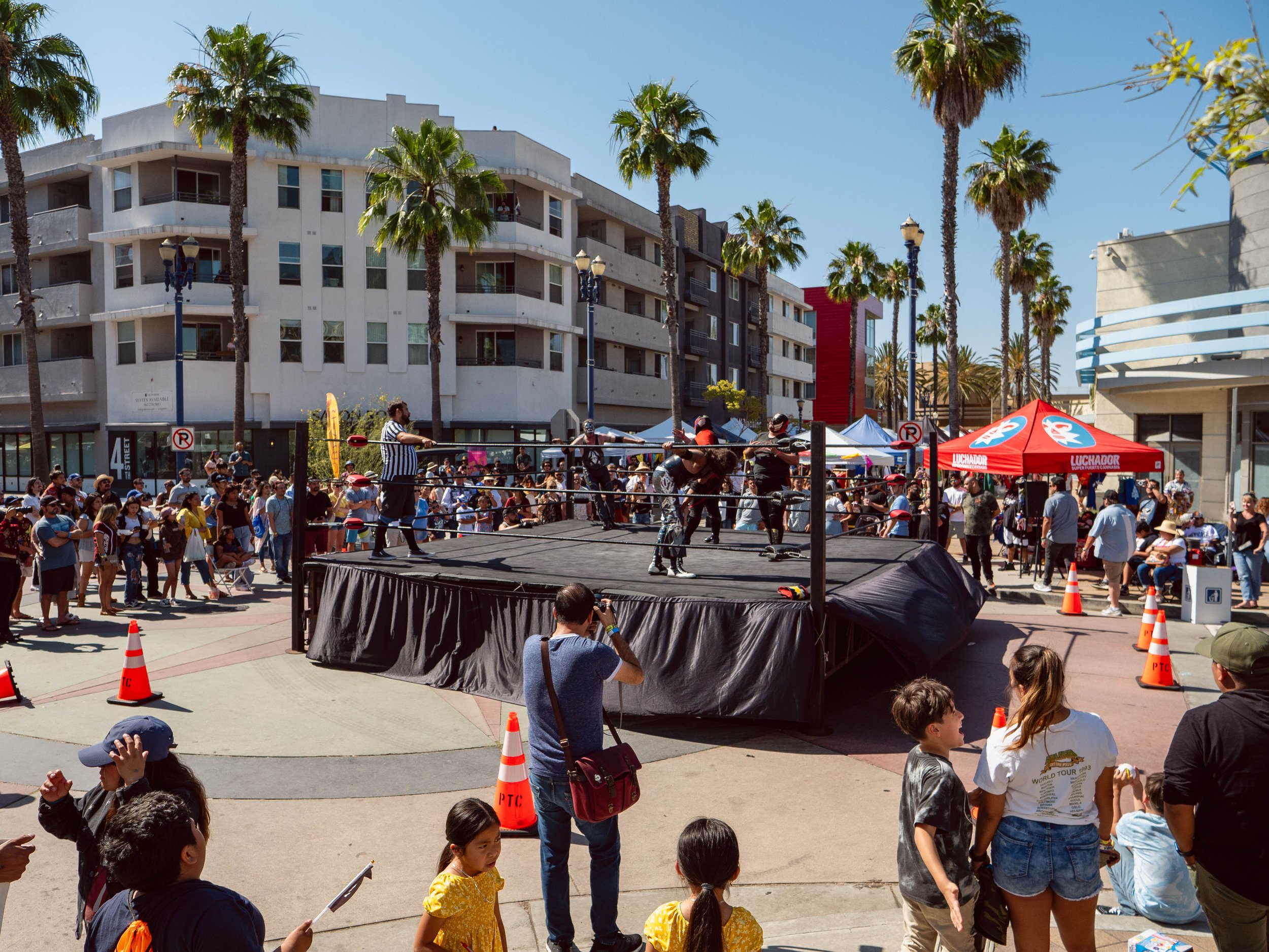 A wrestling match taking place in an outdoor arena with a crowd watching, surrounded by palm trees and buildings on a sunny day.