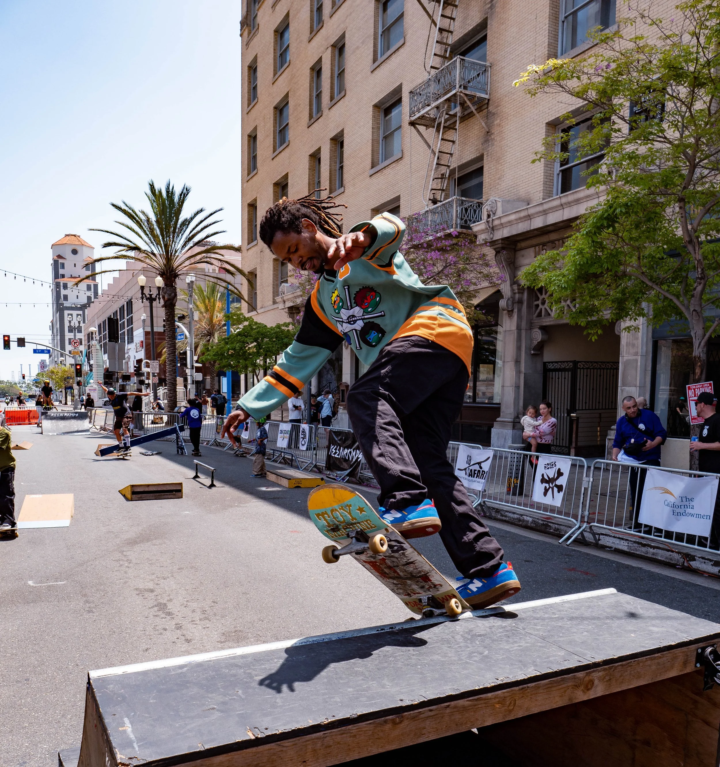 Skateboarder in colorful jacket performing a trick on a ramp in an urban street, with onlookers and buildings in the background.