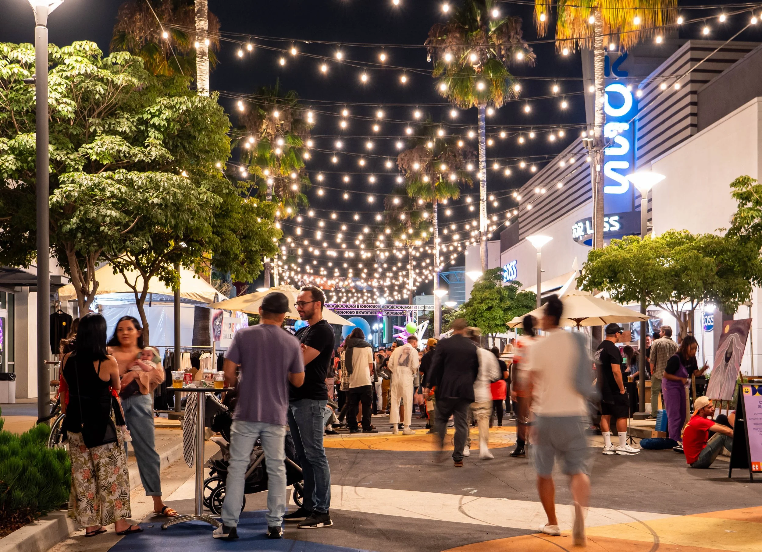 Night scene of a vibrant outdoor shopping or dining area with string lights overhead, several people talking and walking, surrounded by trees, storefronts, and illuminated signs.