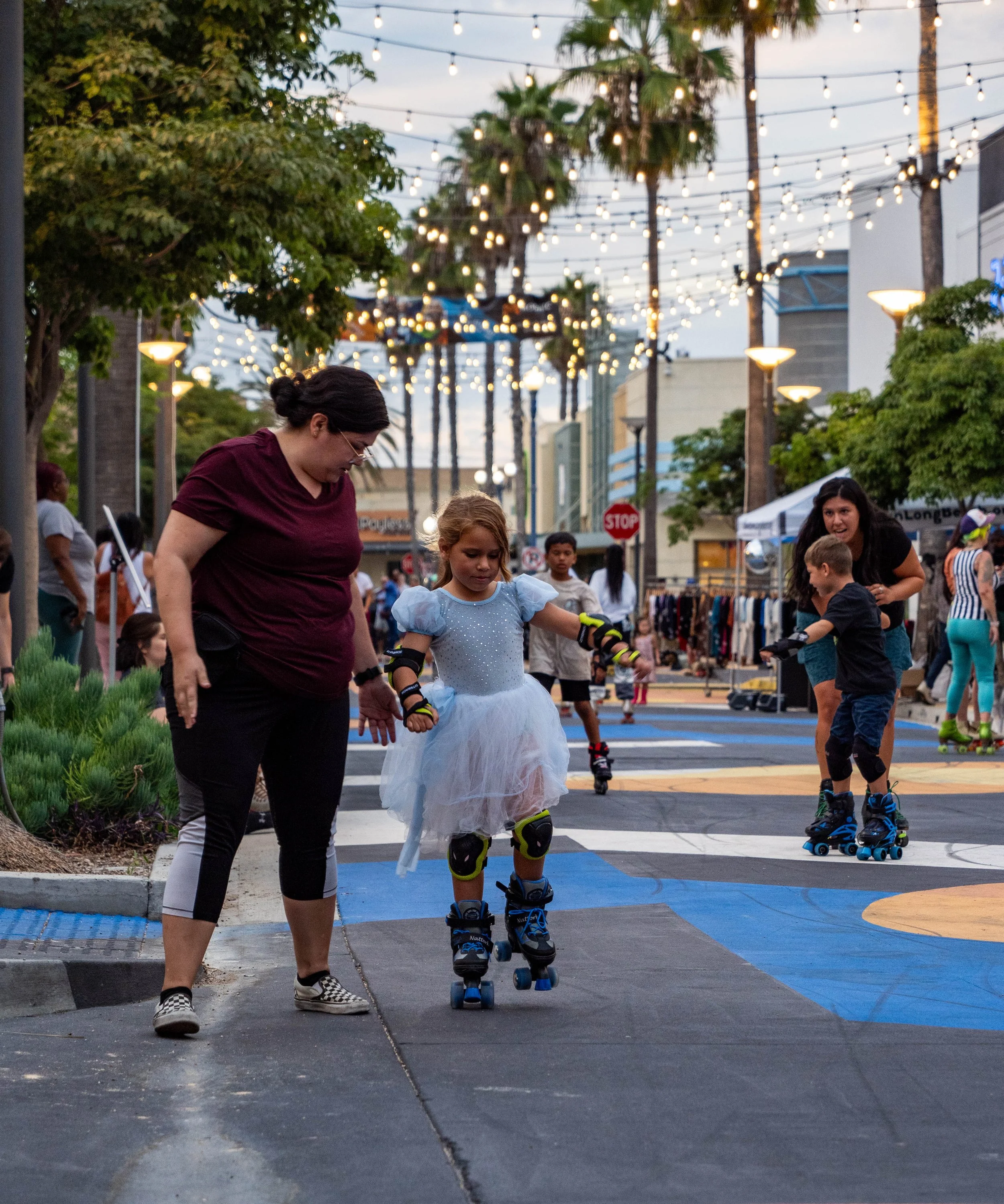 Children roller skating on street at outdoor market with string lights overhead at dusk