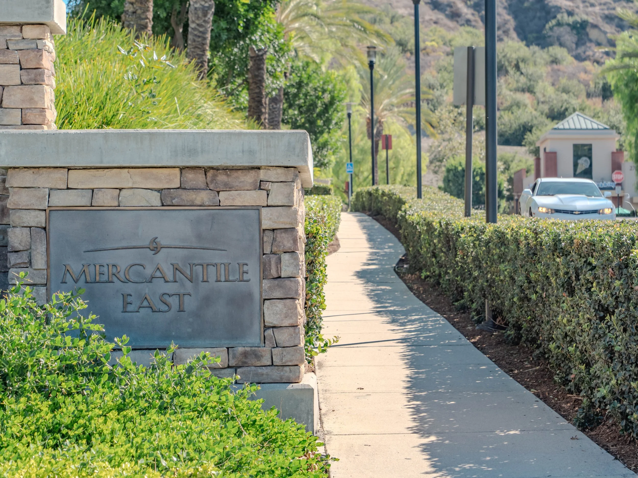 A sidewalk with a stone wall displaying a sign that reads 'Mercantile East' in a landscaped area with bushes, trees, and street lamps, with a road and cars in the background.
