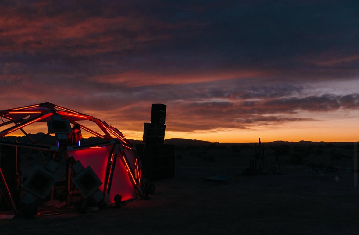 A geodesic dome structure illuminated with red lights on a desert landscape during sunset, with some equipment and scenery in the background.